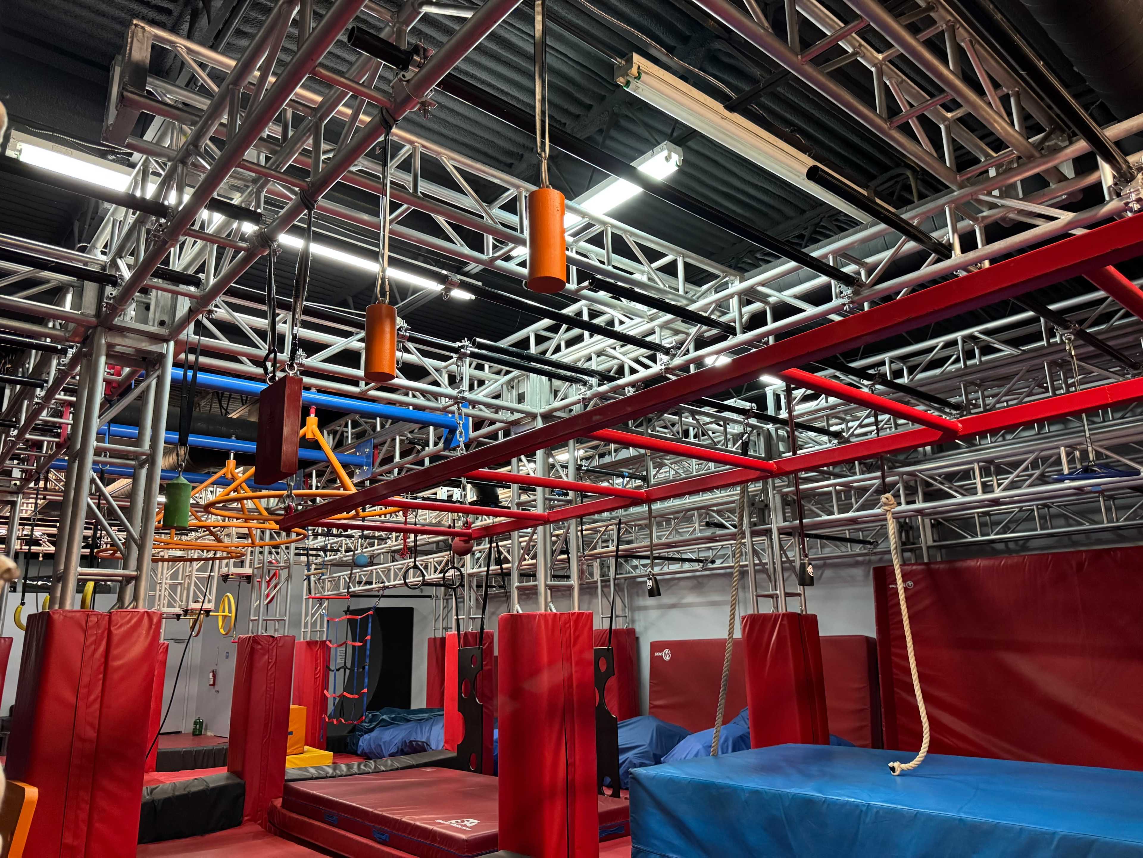 The image shows an indoor obstacle course with metal frames, hanging apparatuses, and padded areas for training or exercise.