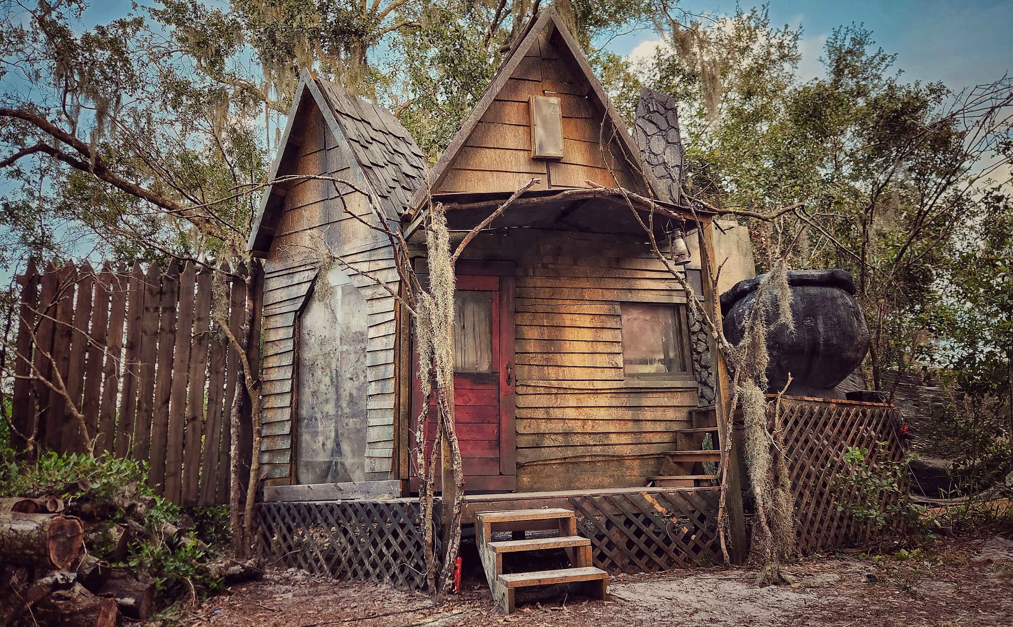 A rustic, two-story wooden house in a wooded area, featuring a steeply pitched roof and a red door.