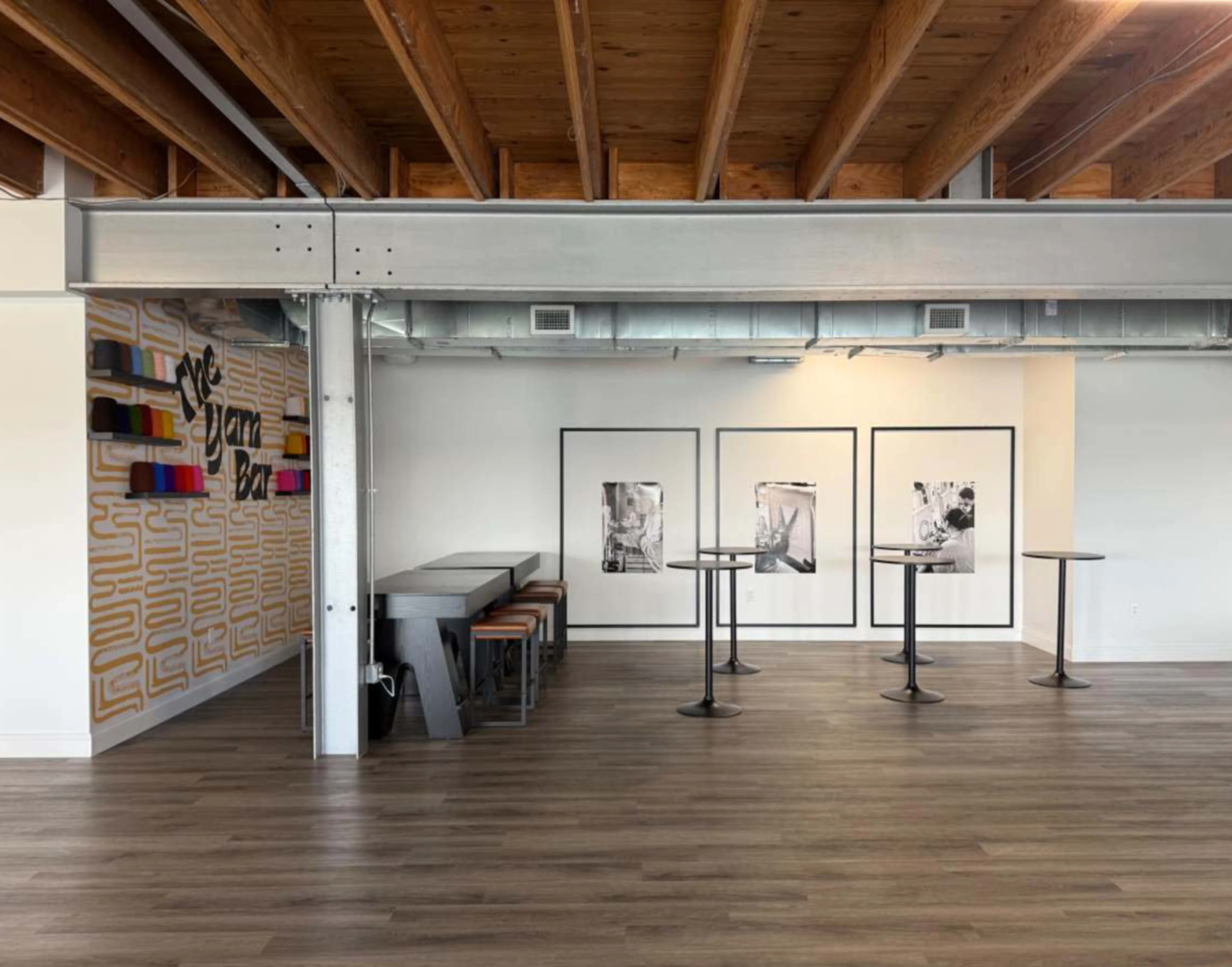 The image shows a spacious interior with wooden beams and flooring, featuring a seating area with high tables and art displayed on the walls.