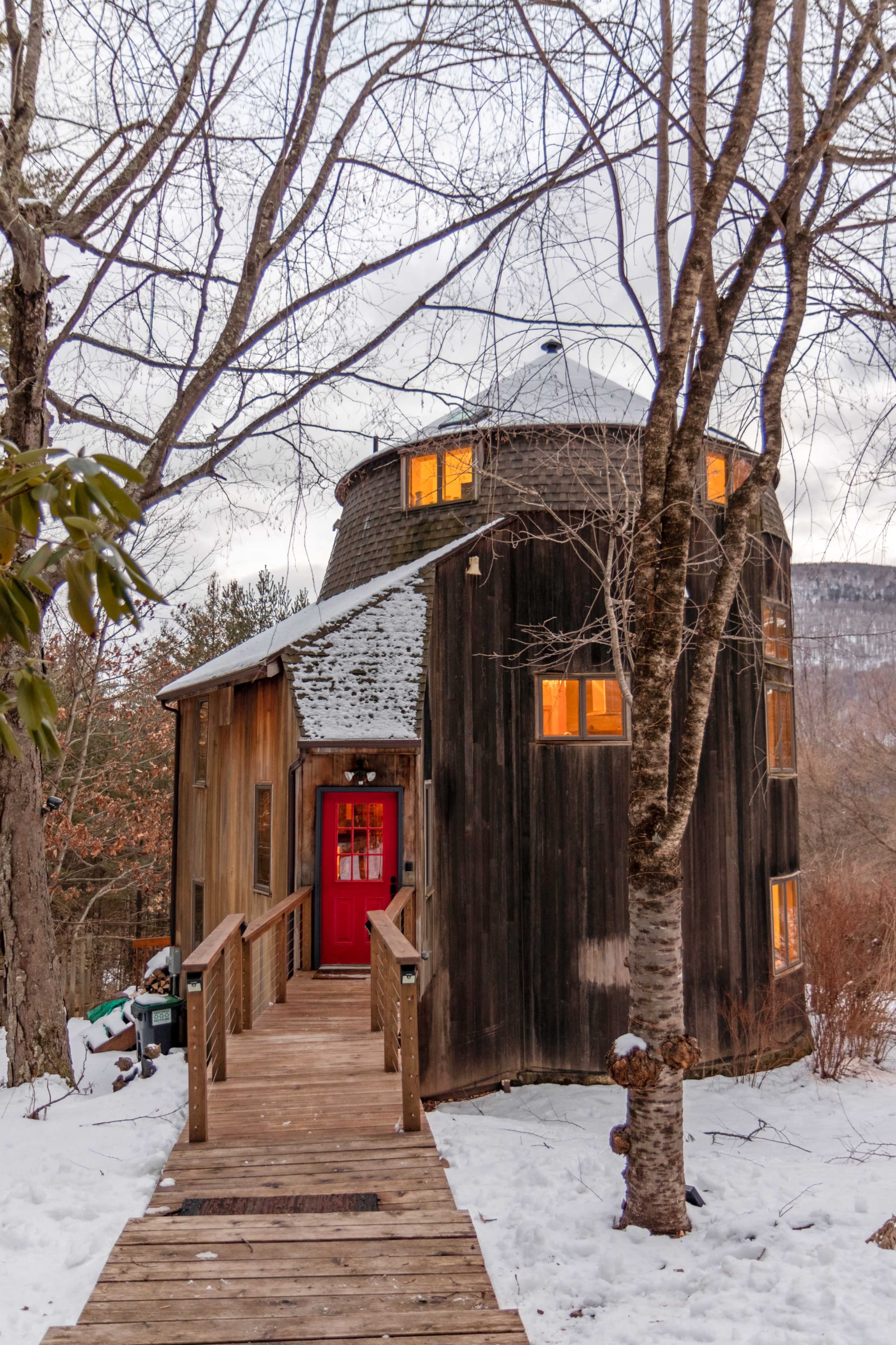 A unique round wooden house with a red door is set against a snowy landscape, surrounded by bare trees.