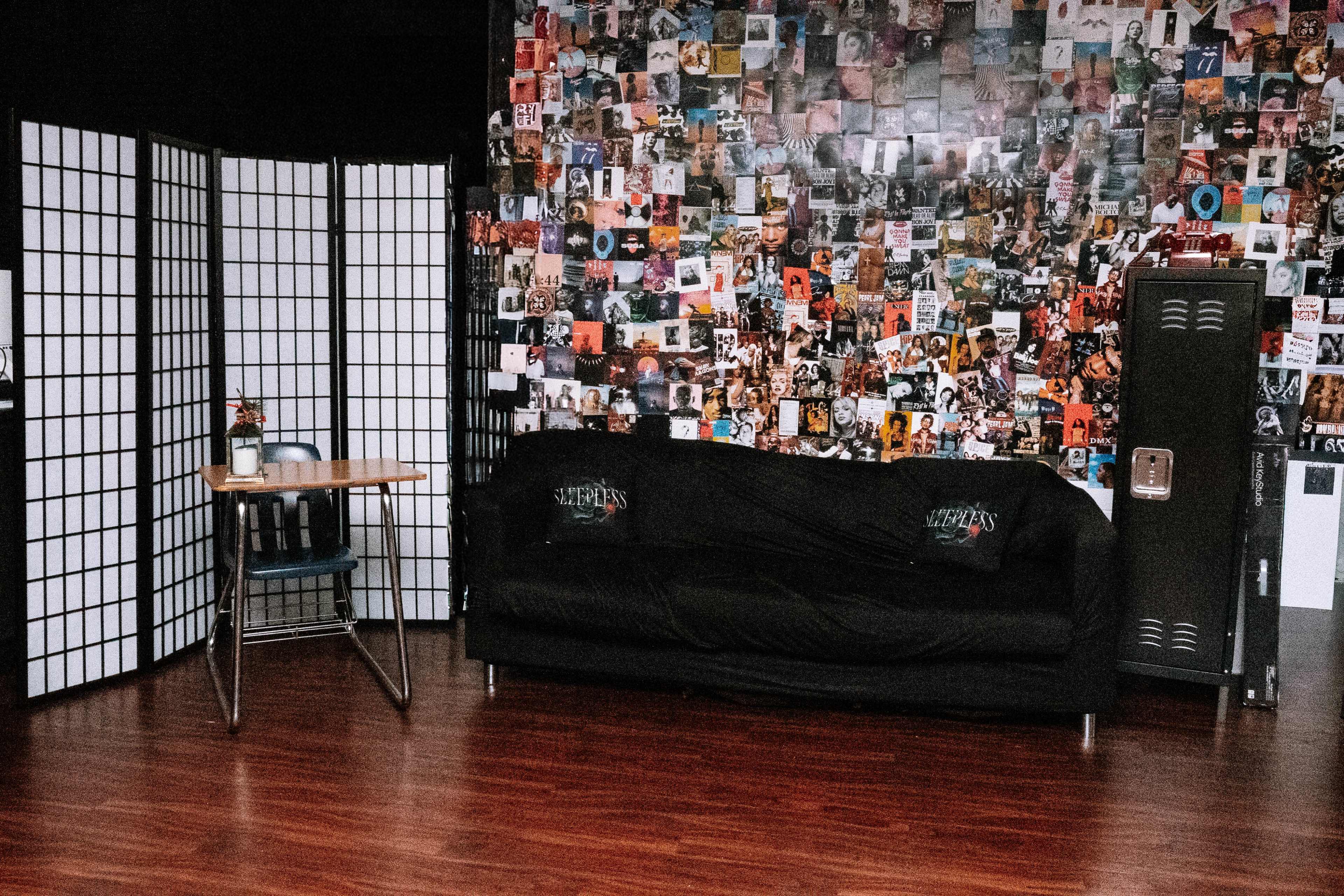 The image shows a small room with a black couch, a table with drinks, a folding screen, and a wall covered in various posters.
