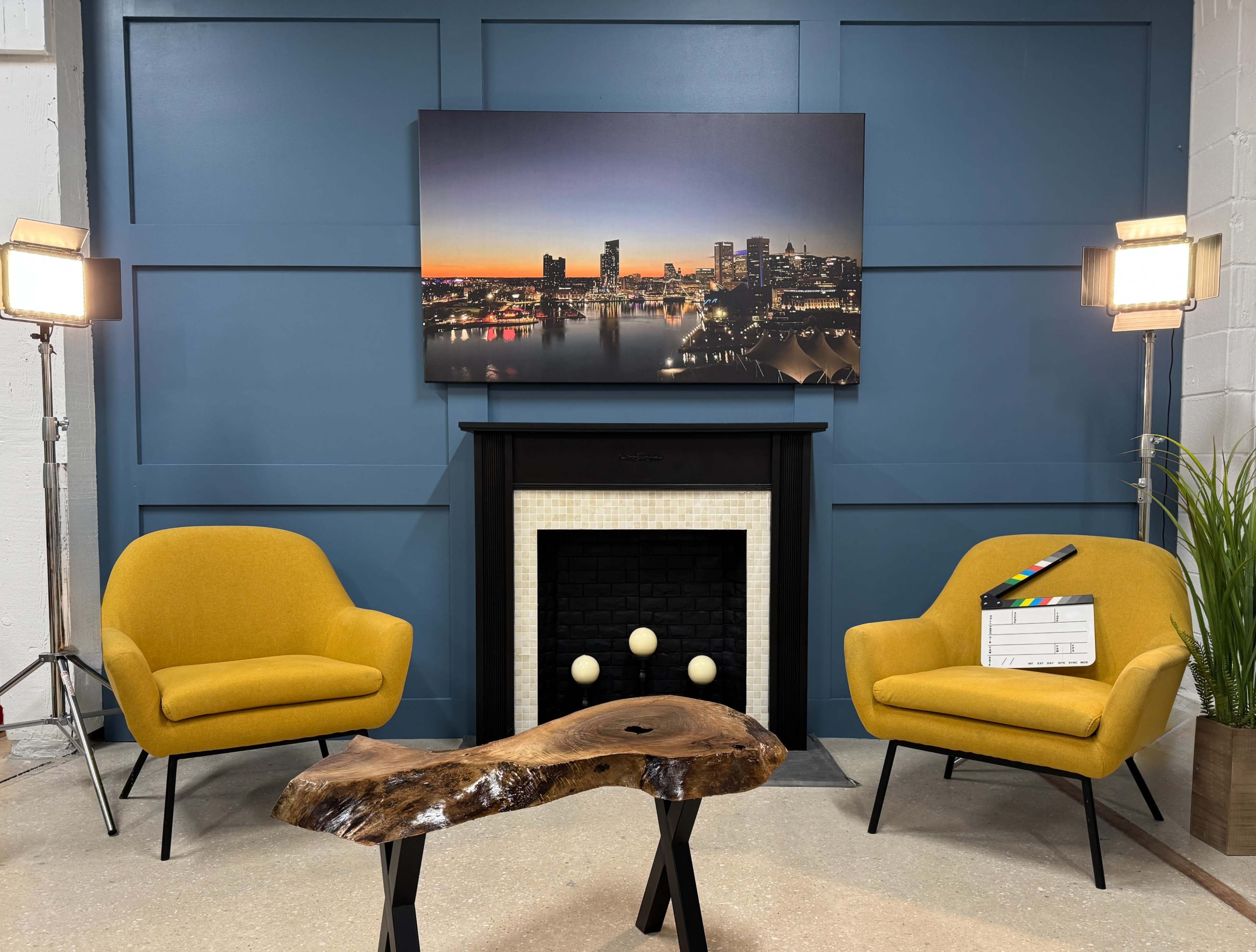 The image shows a cozy studio setup with two yellow armchairs facing a black fireplace, a large photographic print of a city skyline at sunset on the wall, and a rustic wooden table in the center.