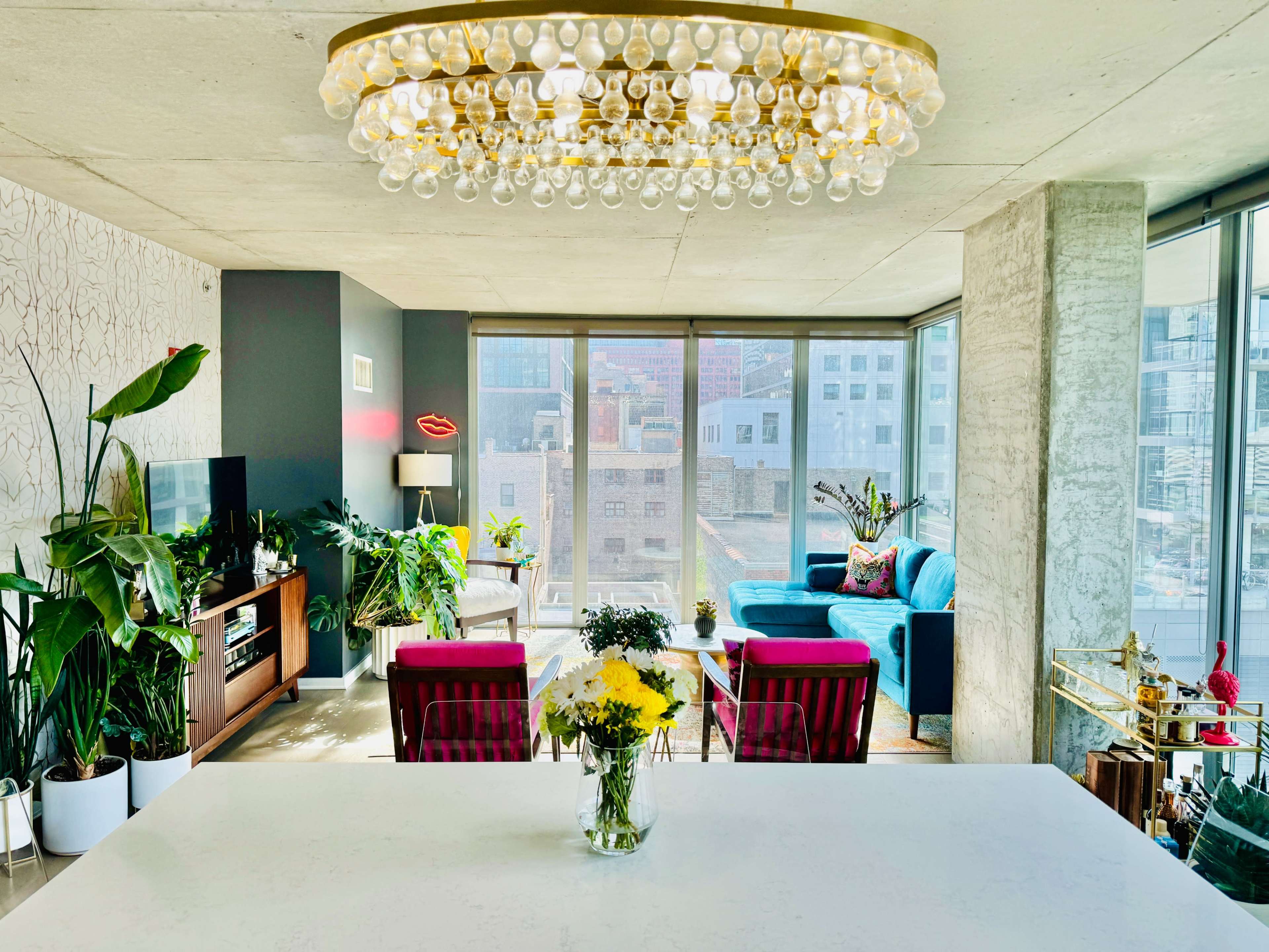 A modern living room features large windows with a city view, a decorative chandelier, colorful furniture, and indoor plants.