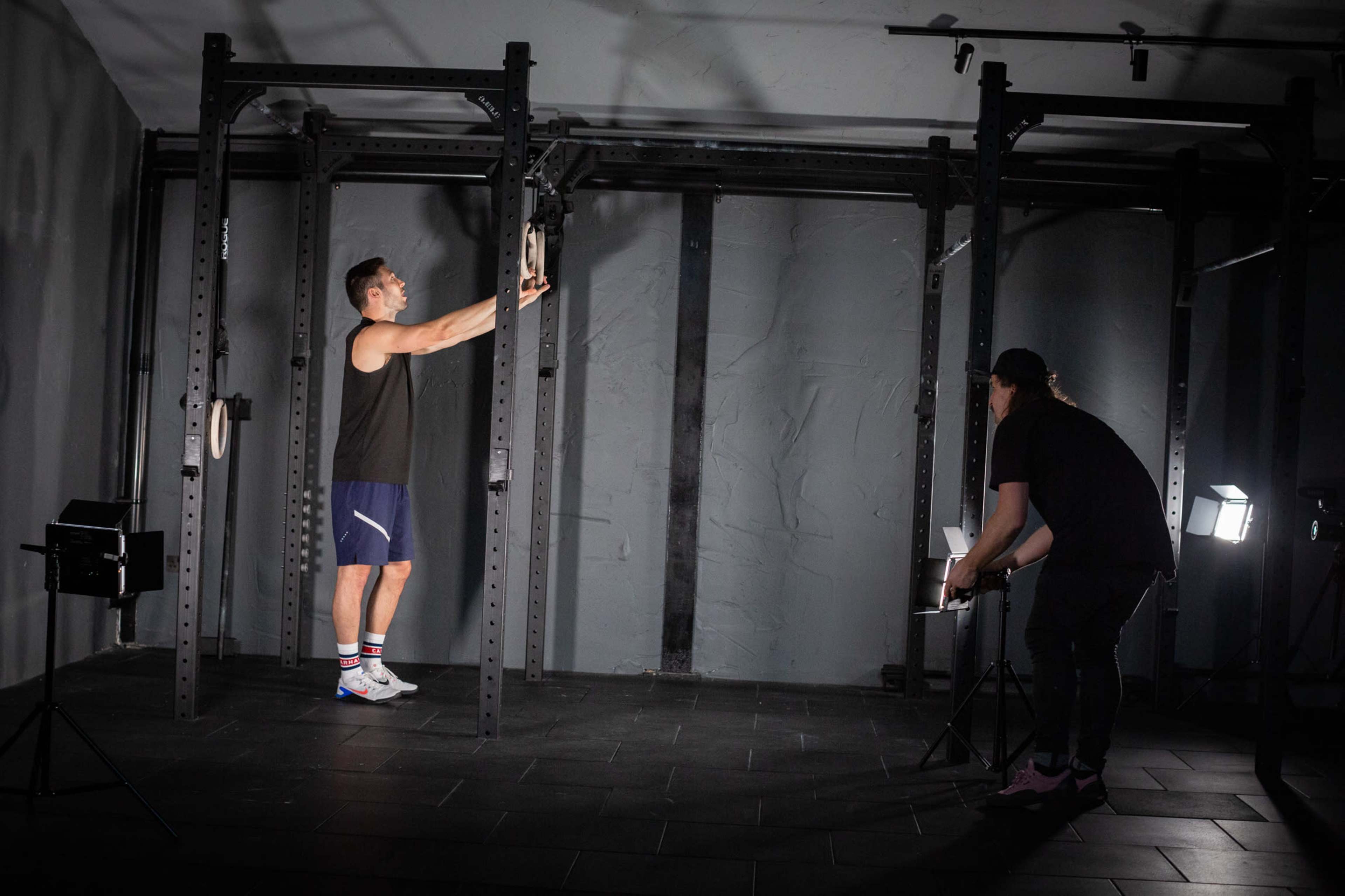 A person performs a workout in a gym while another individual adjusts lighting equipment nearby.