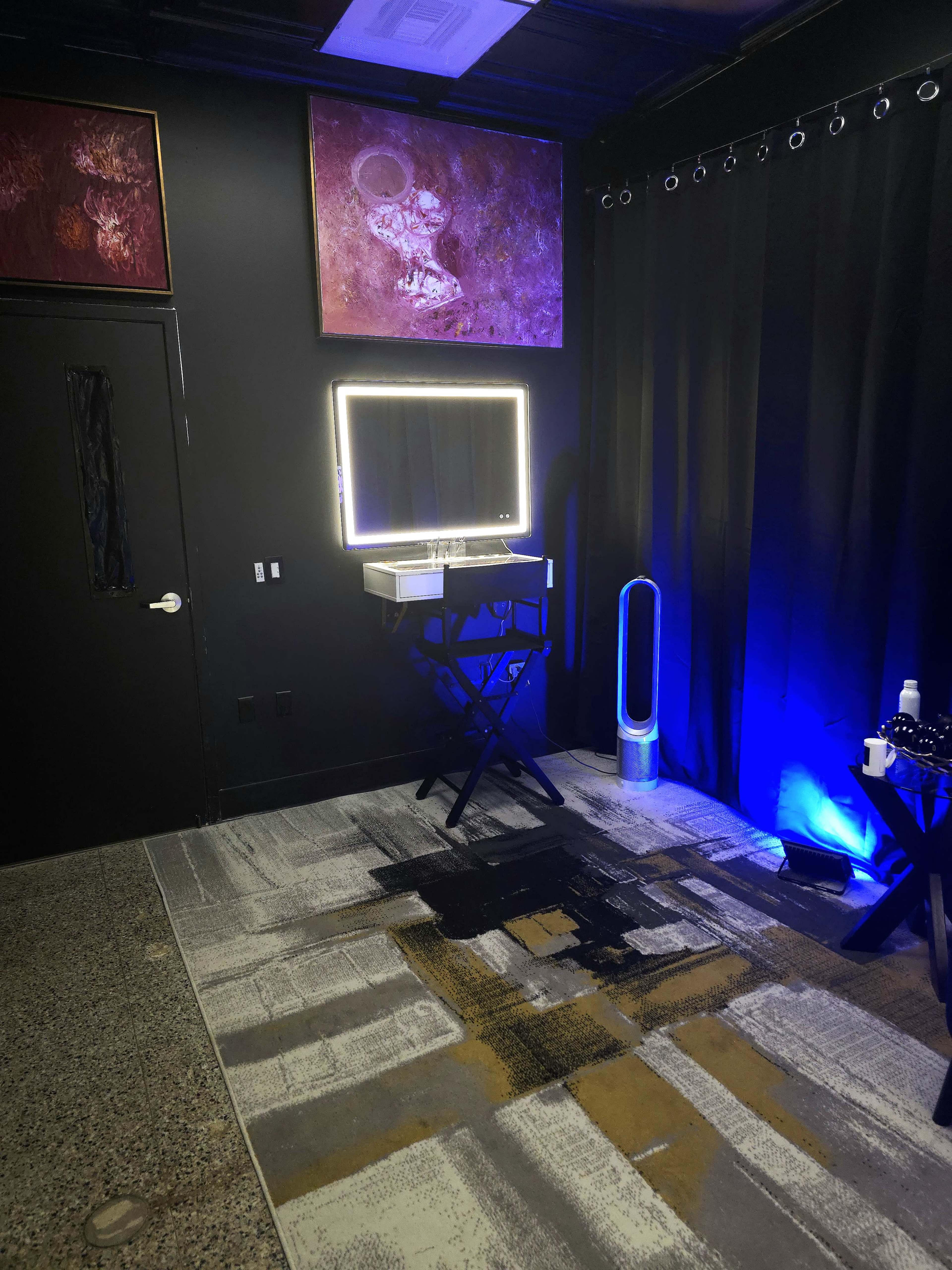 The image shows a dimly lit room with a black wall, featuring a mirror on a stand, a decorative rug, and artwork hanging on the walls.