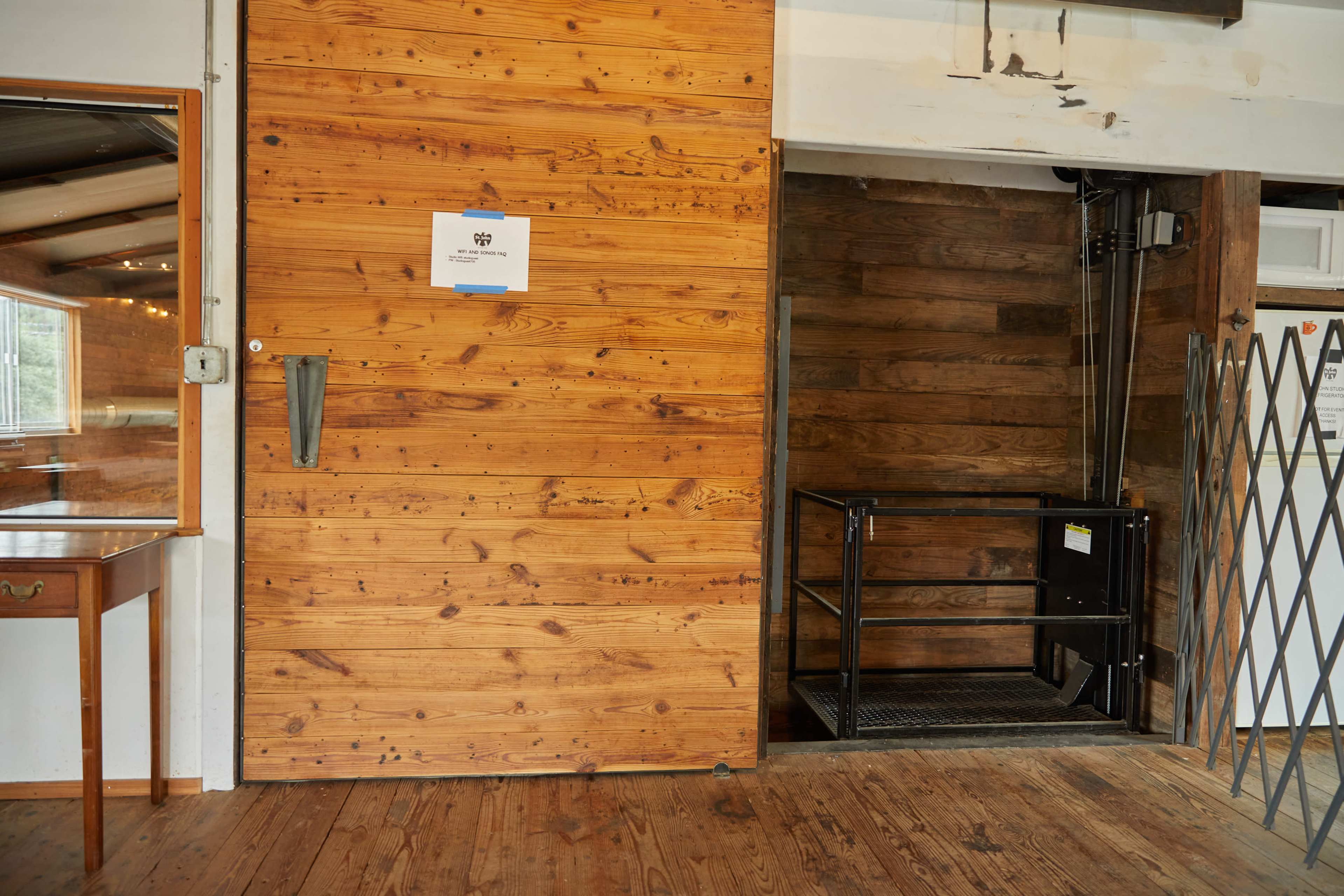 A wooden interior with a large, textured door on the left and a small freight elevator with a black platform on the right.