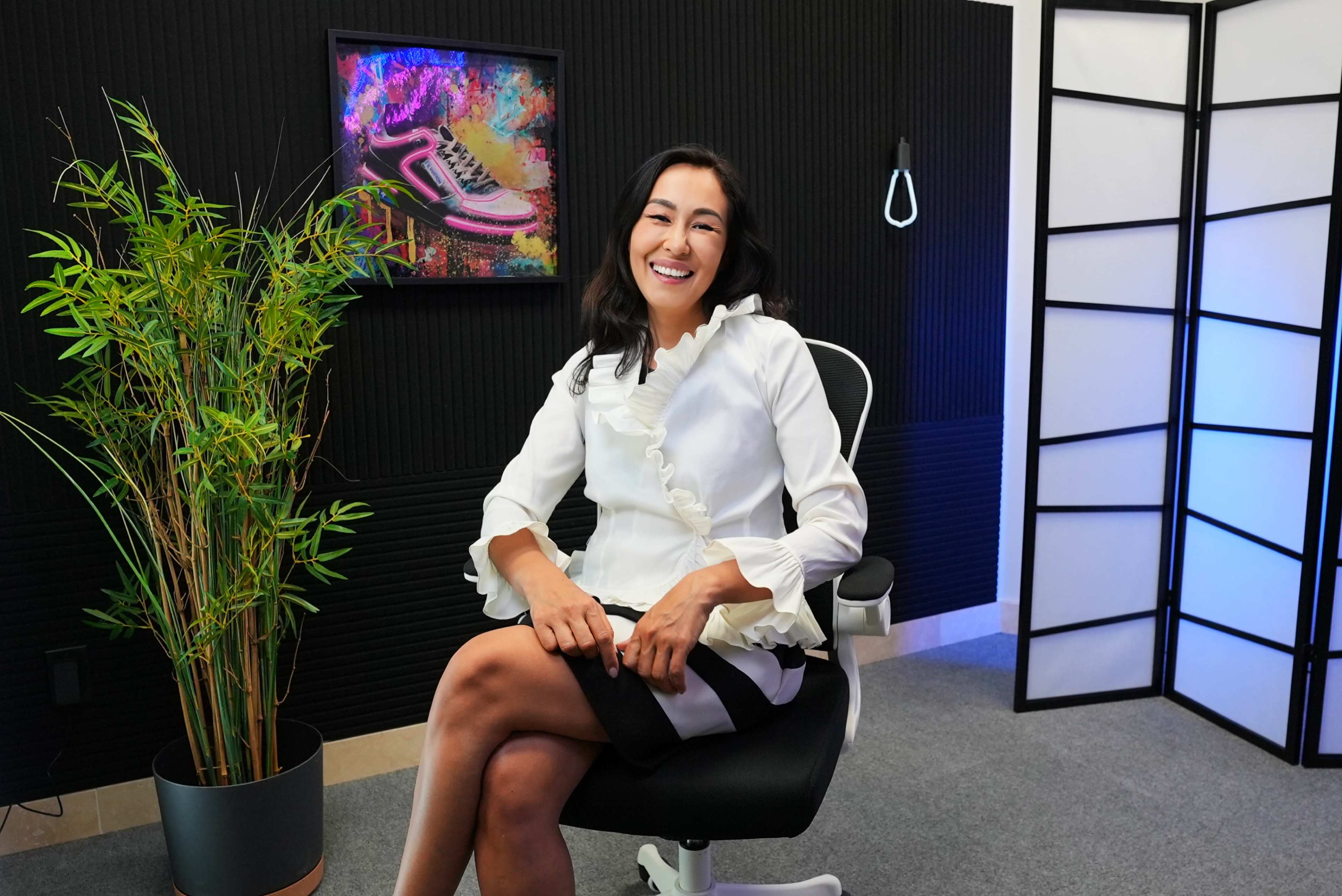 A woman in a white blouse sits in an office chair, smiling, with a plant and a colorful framed artwork in the background.