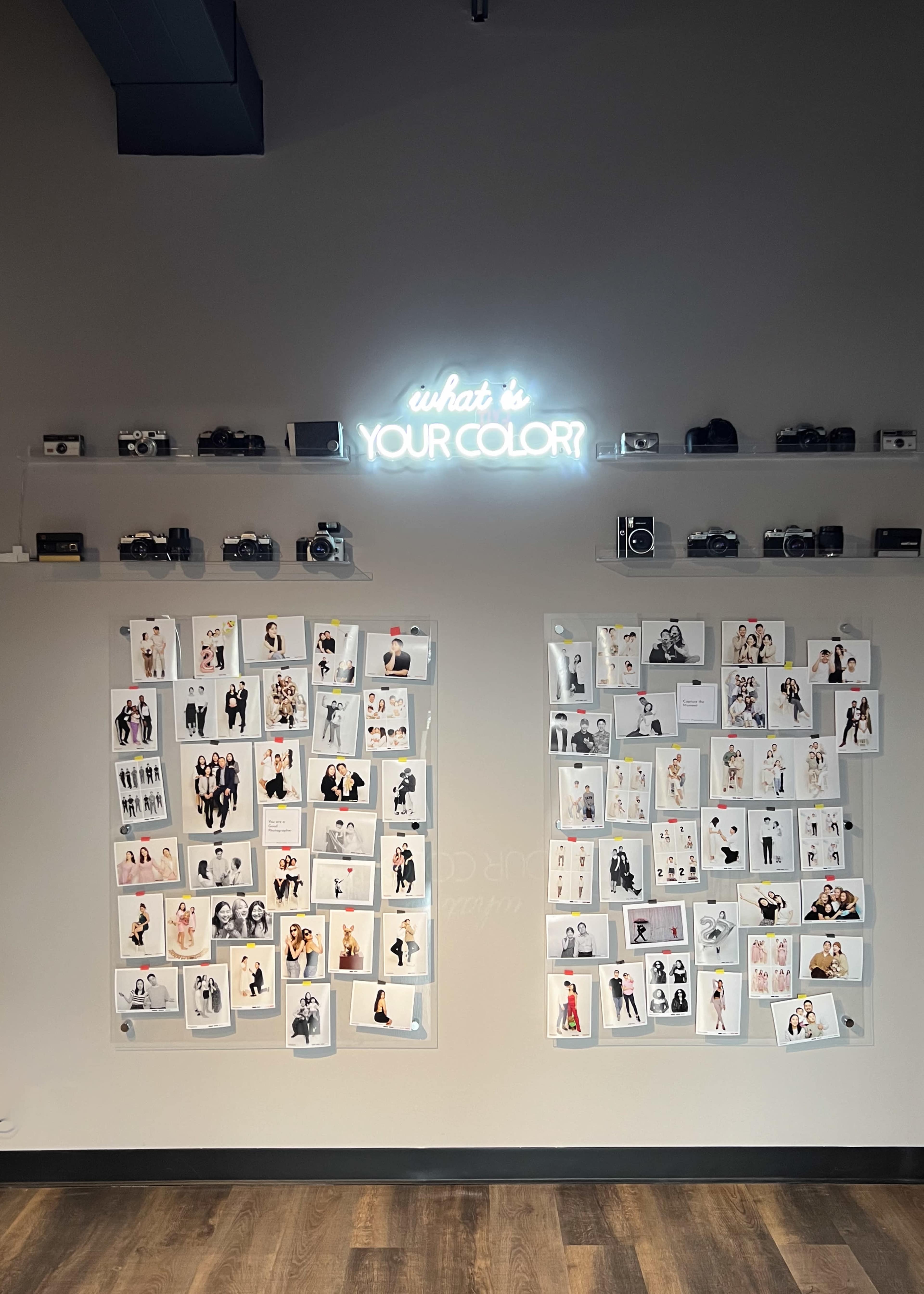 The image shows a wall with several photographs arranged in a grid, alongside a neon sign that reads "what is YOUR COLOR?" and a shelf displaying vintage cameras above.