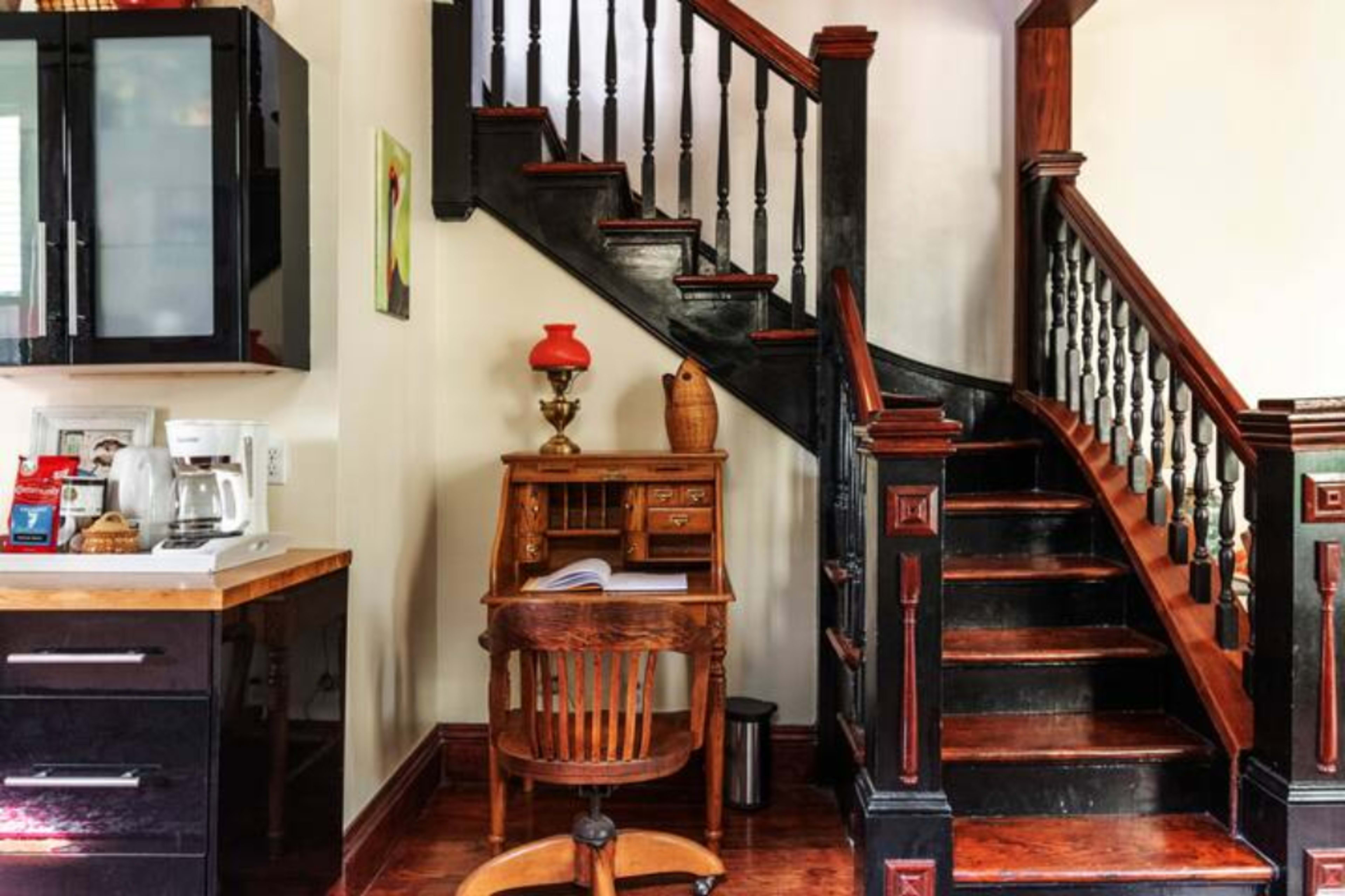 The image shows a quaint interior featuring a wooden staircase, a small desk with a chair, and a kitchen area with a coffee maker.
