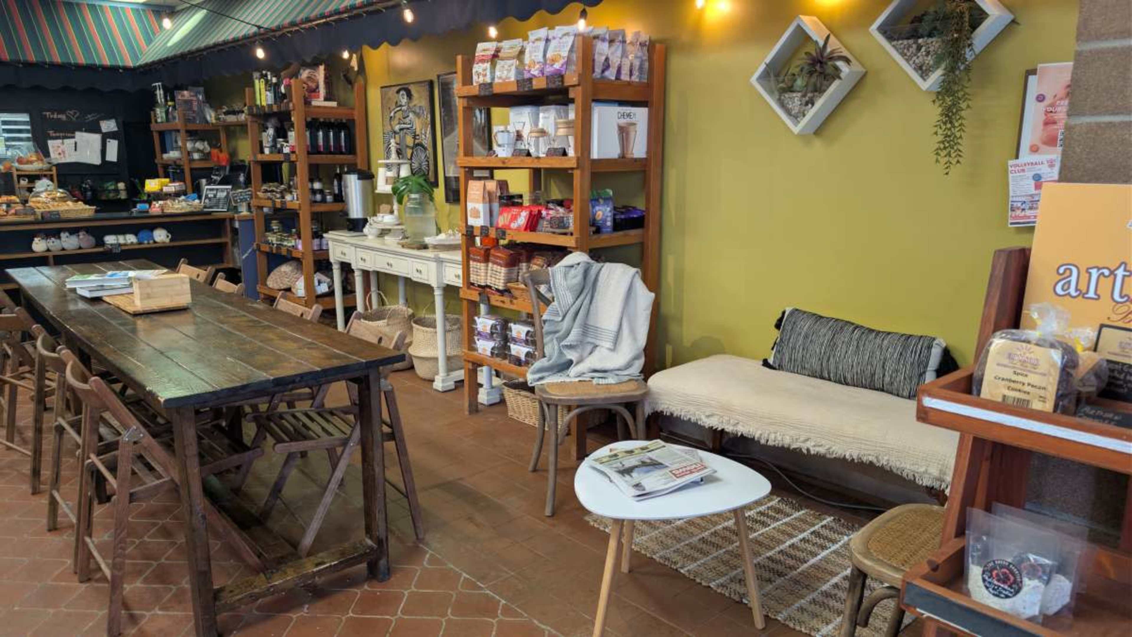 The image shows a cozy interior of a café or shop featuring wooden shelves filled with various products, a long table with chairs, and a small seating area with a sofa and coffee table.
