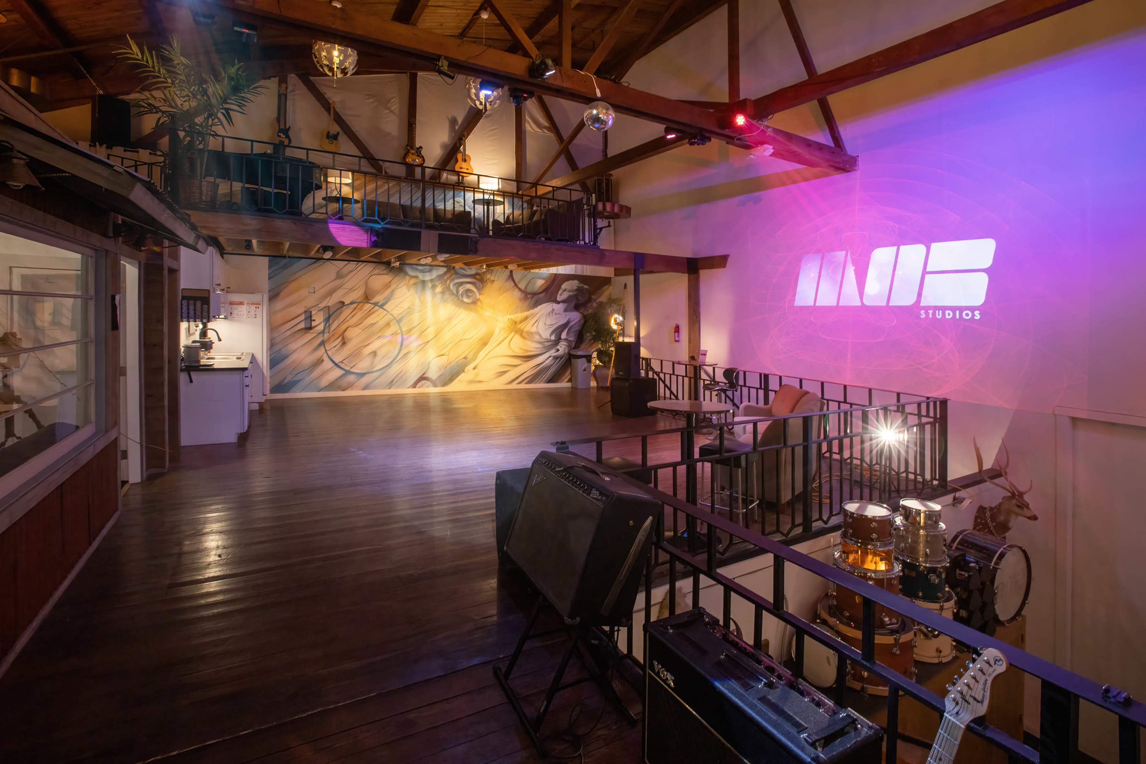 A spacious studio with wooden beams, a mural on the wall, musical instruments set up, and dim lighting featuring colored spotlights.