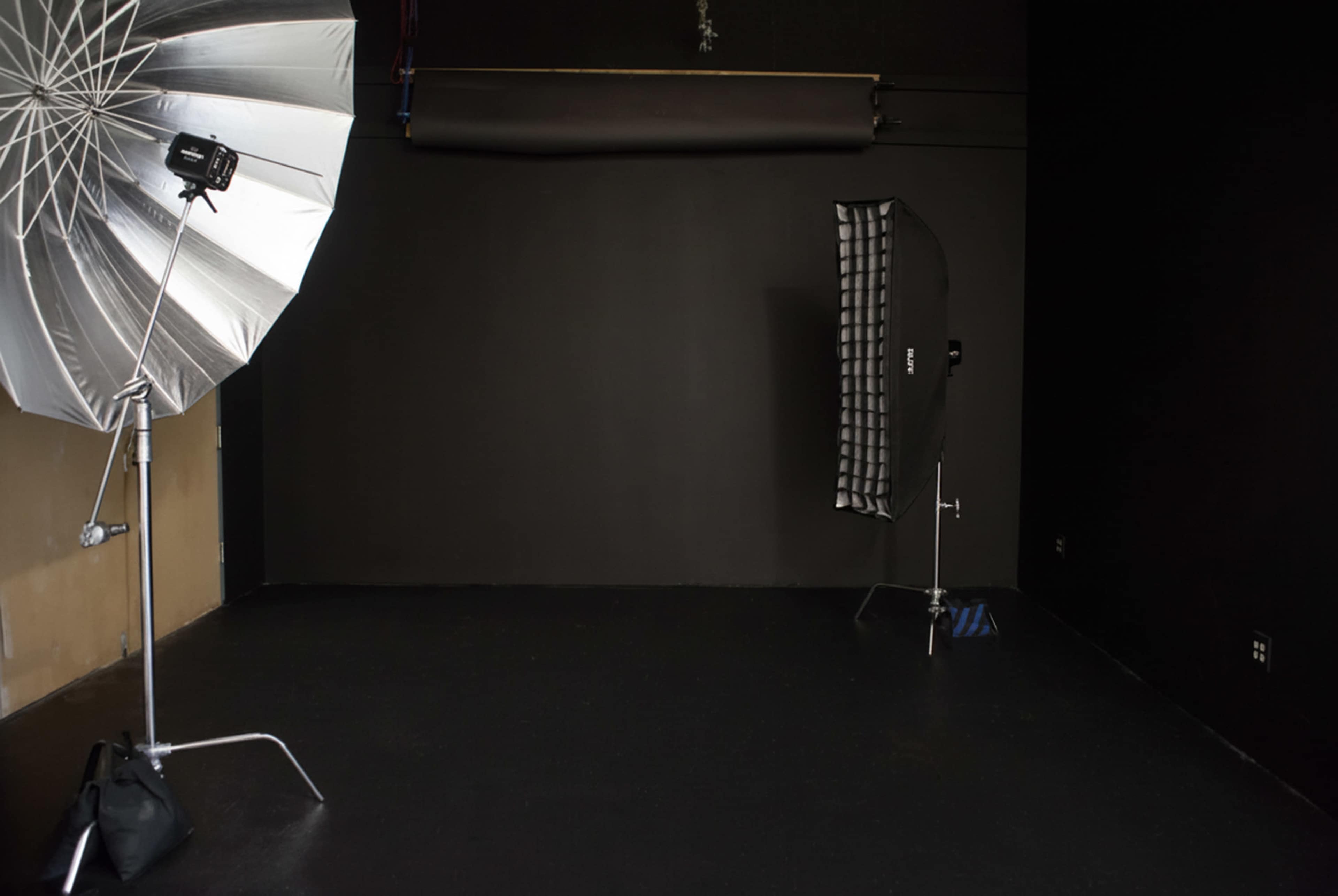 The image shows a photography studio with a large umbrella light on one side and a softbox on the other, set against a black backdrop and floor.