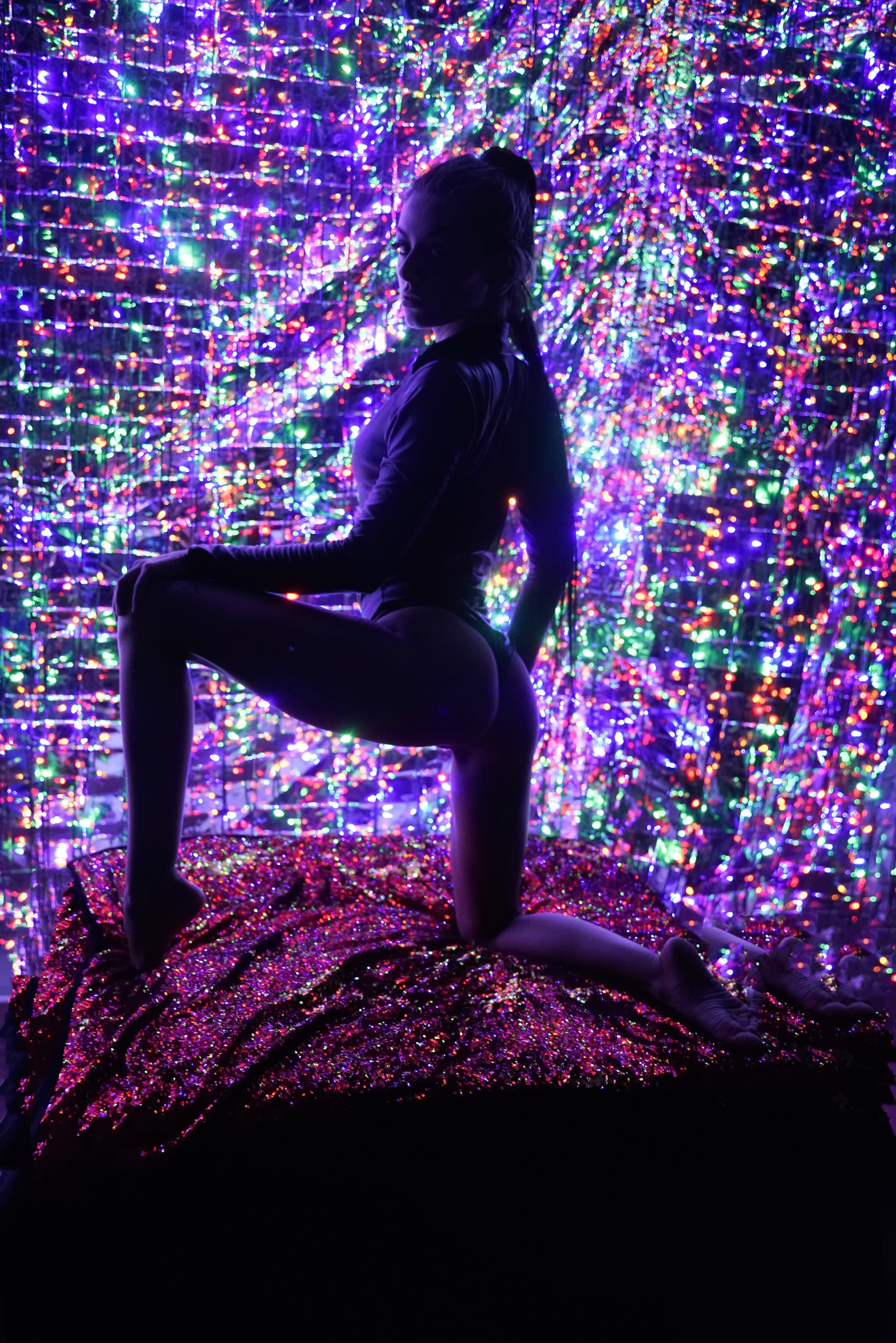 A person poses on a textured surface in front of a vibrant, multicolored sequin backdrop.