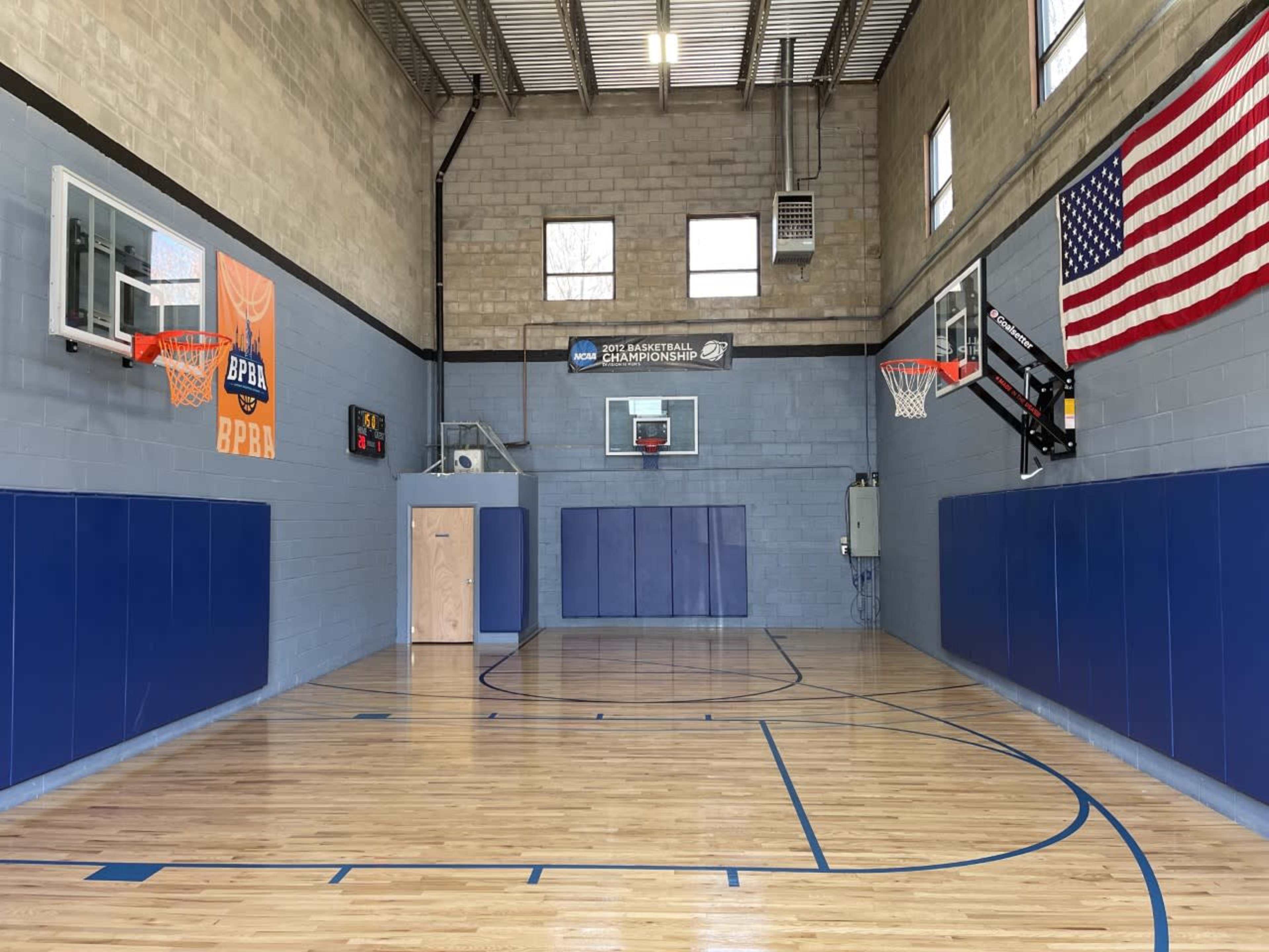An indoor basketball court featuring two hoops, blue wall padding, and an American flag on the wall.