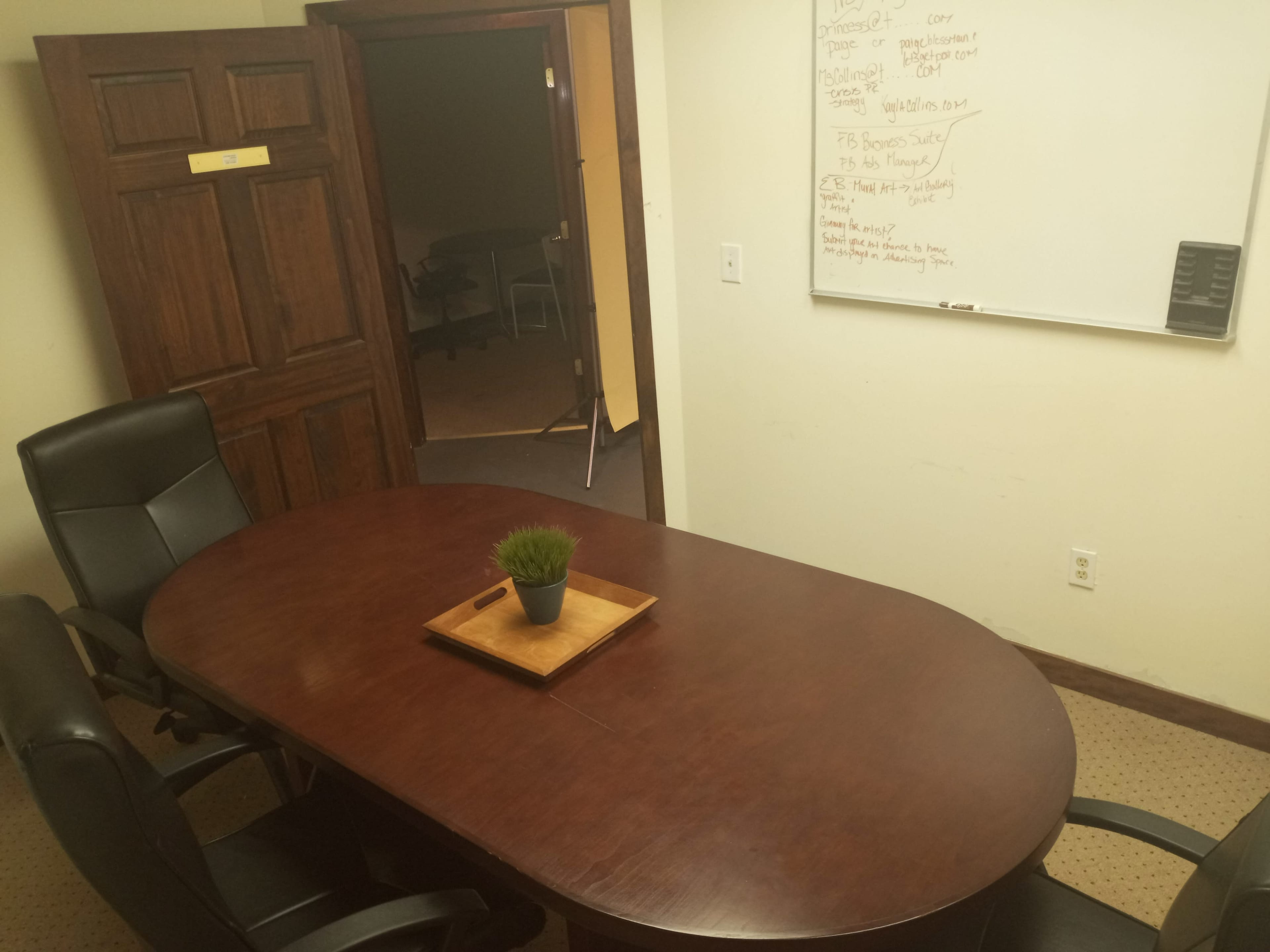 A conference room features a large wooden table surrounded by black leather chairs, with a small potted plant on the table and a whiteboard on the wall.