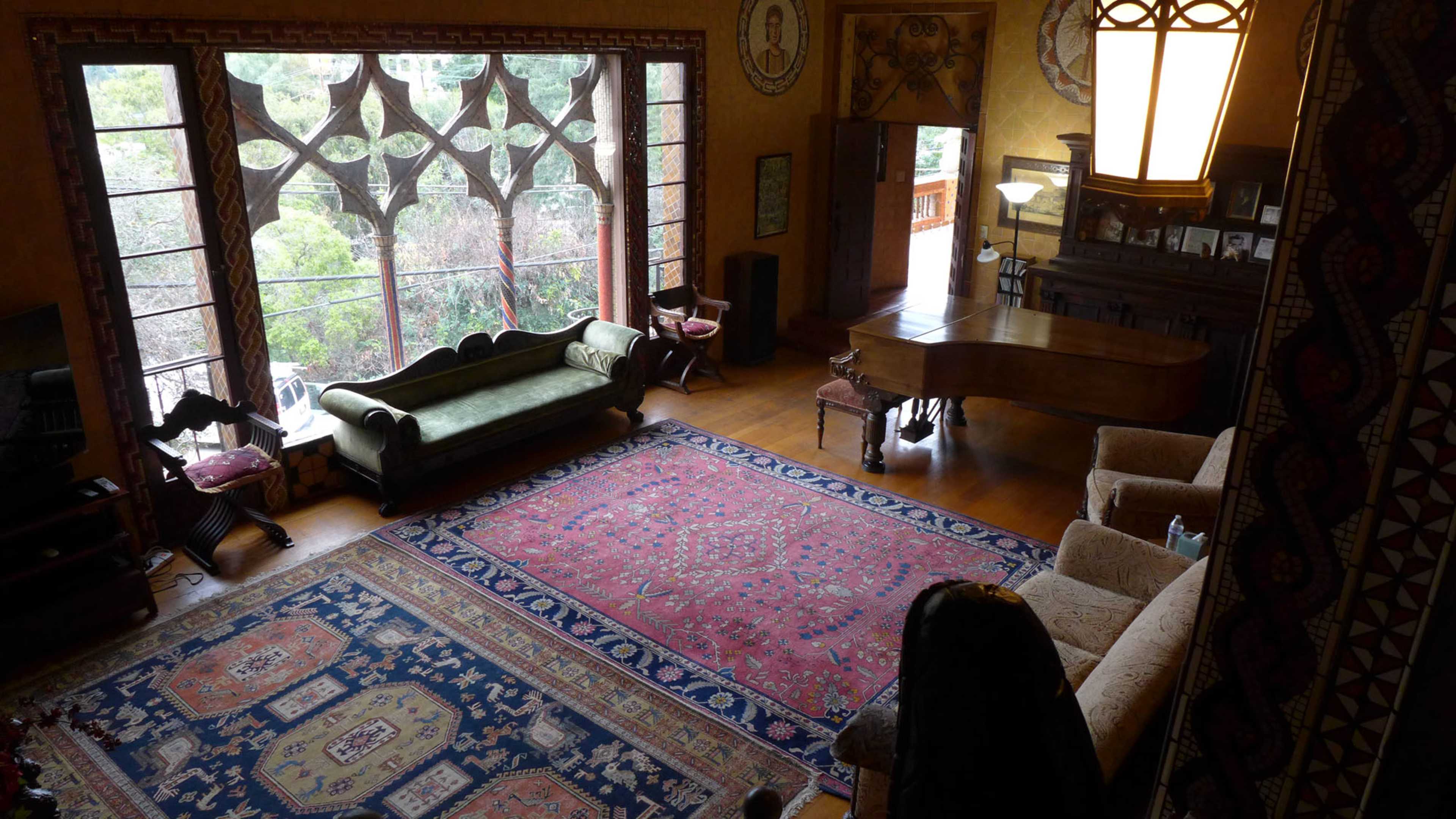 A spacious living room features large windows, a grand piano, and a patterned rug surrounded by various seating arrangements.