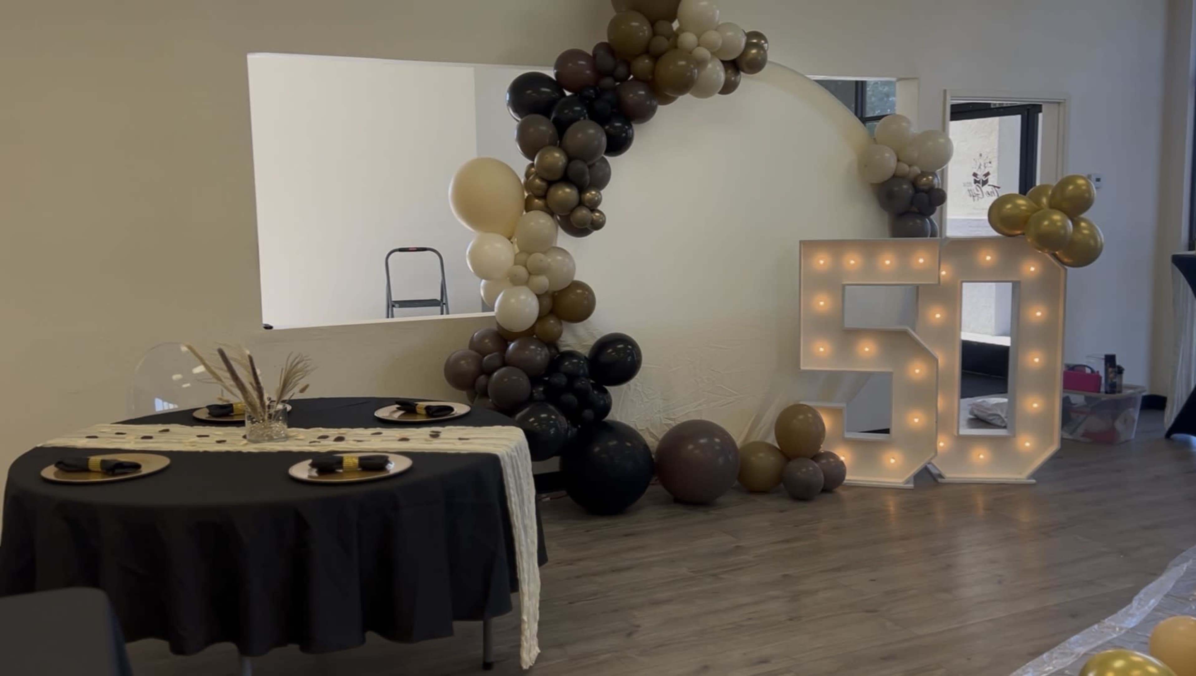 The image shows a decorated indoor space with a large "50" illuminated number, an arch made of balloons in various colors, and two black tables set with plates and napkins.