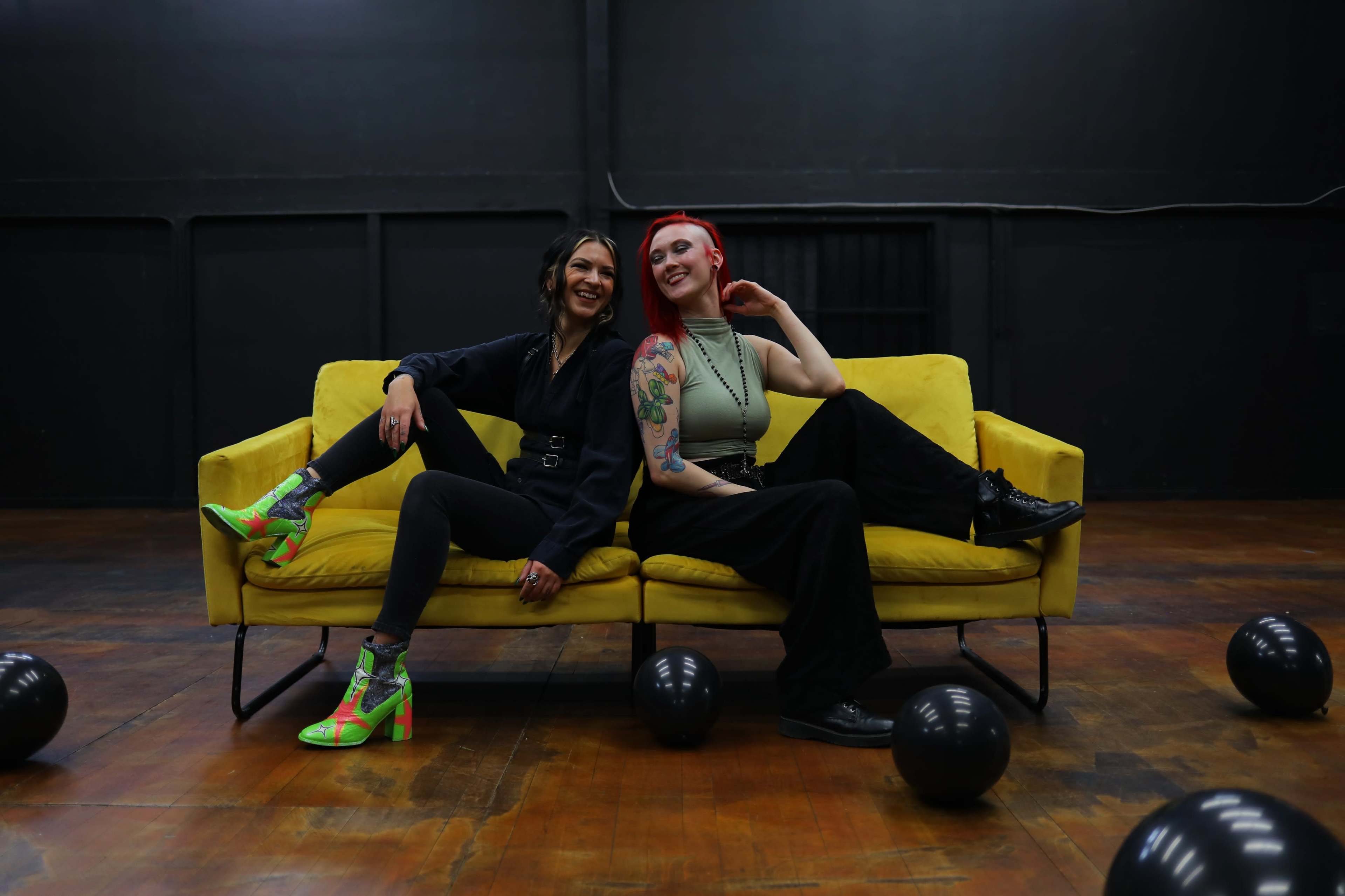 Two women sit on a yellow couch in a dimly lit room with black balloons scattered on the floor.