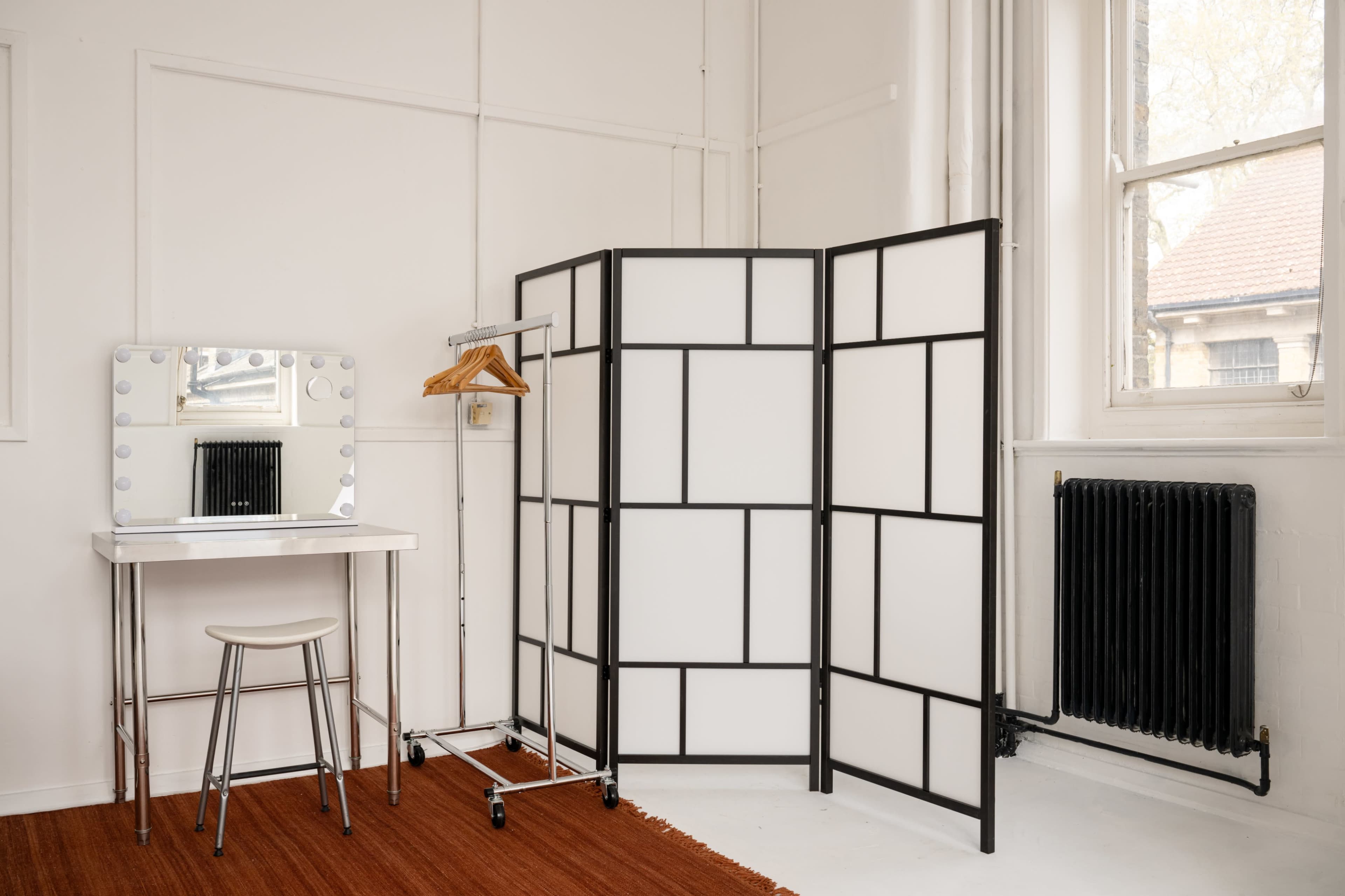 The image shows a minimalist dressing area featuring a vanity mirror with lights, a stool, a clothing rack, and a folding room divider in a bright room with large windows.