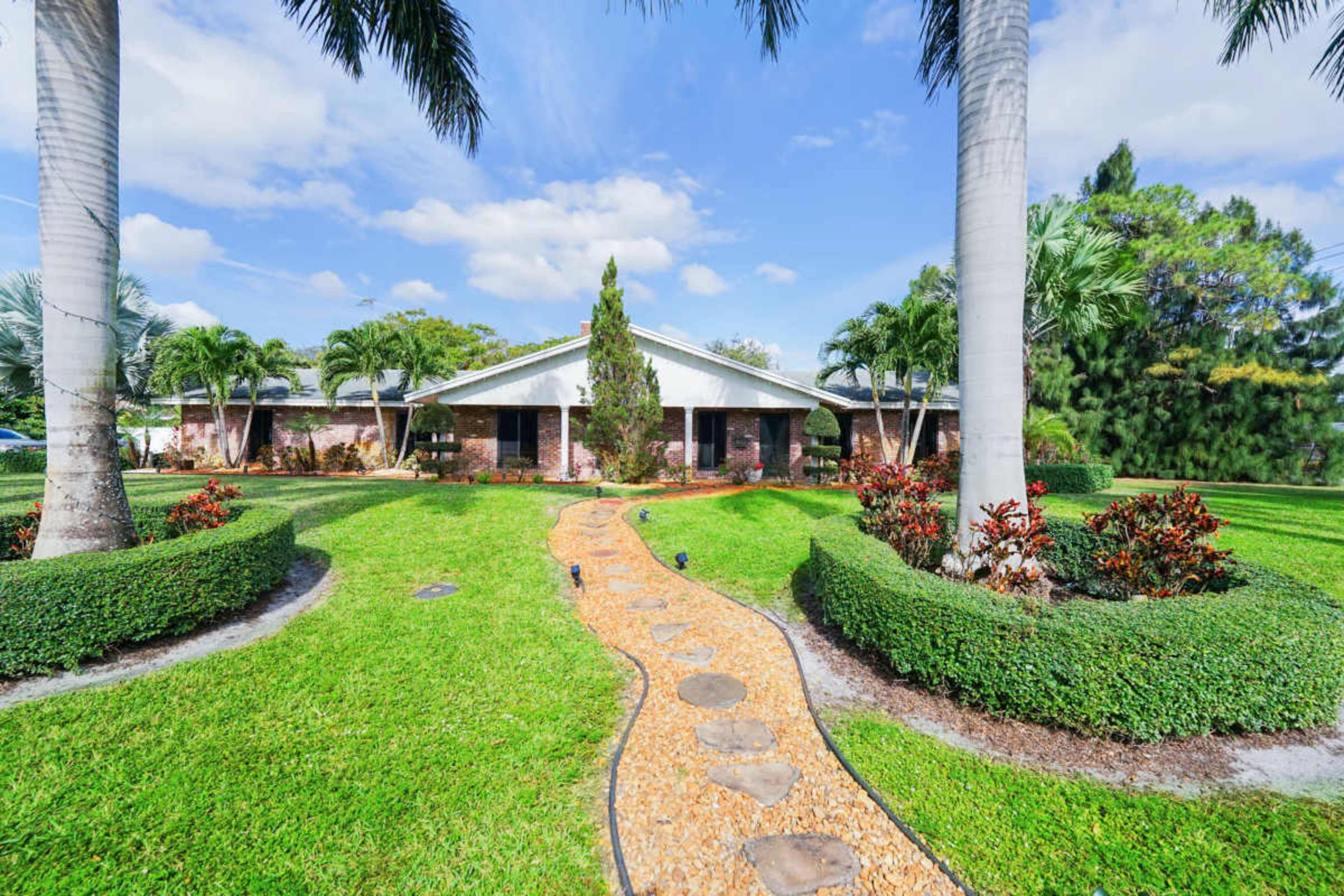 A landscaped yard features a winding stone pathway leading up to a single-story brick house surrounded by palm trees and shrubs.