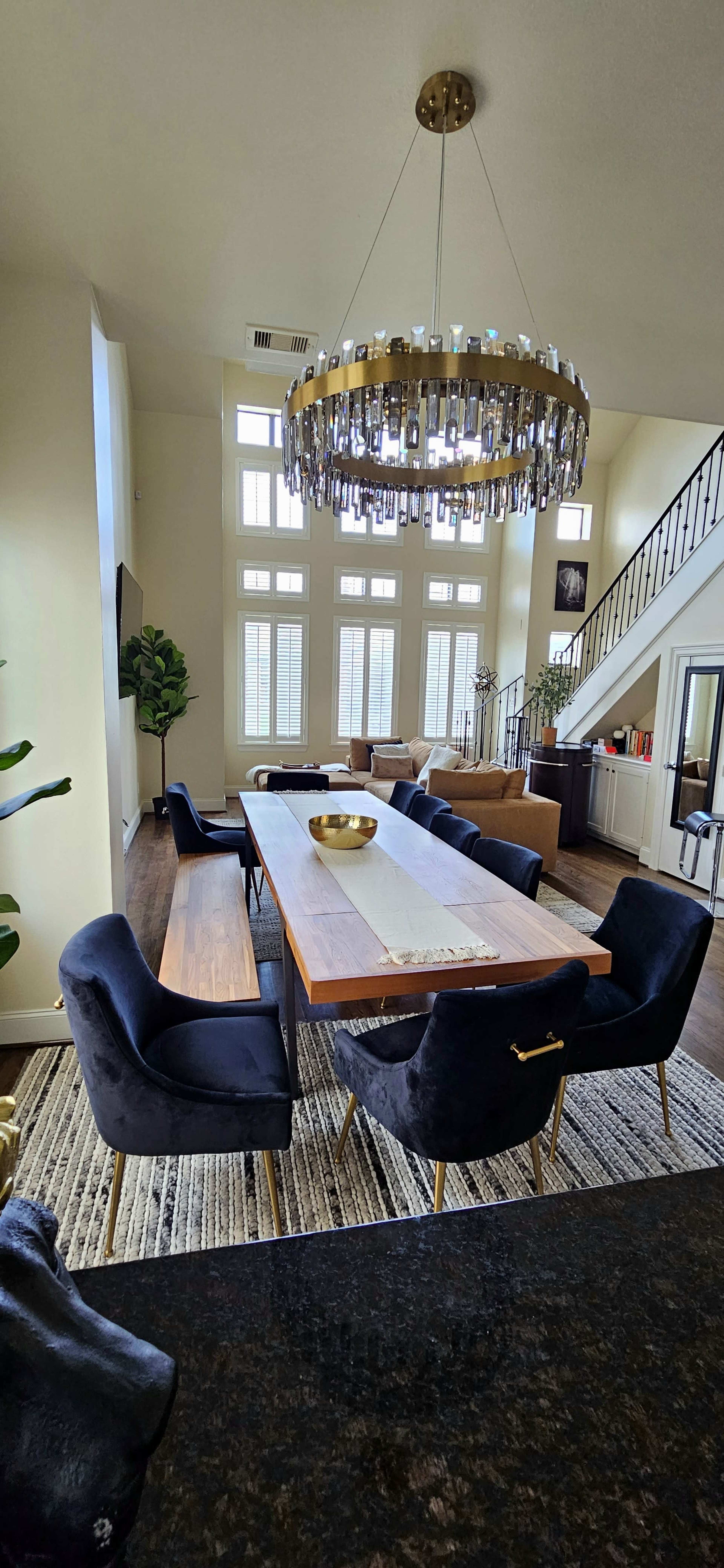 A modern dining room with a long wooden table surrounded by dark upholstered chairs, a large chandelier overhead, and tall windows allowing natural light to enter.