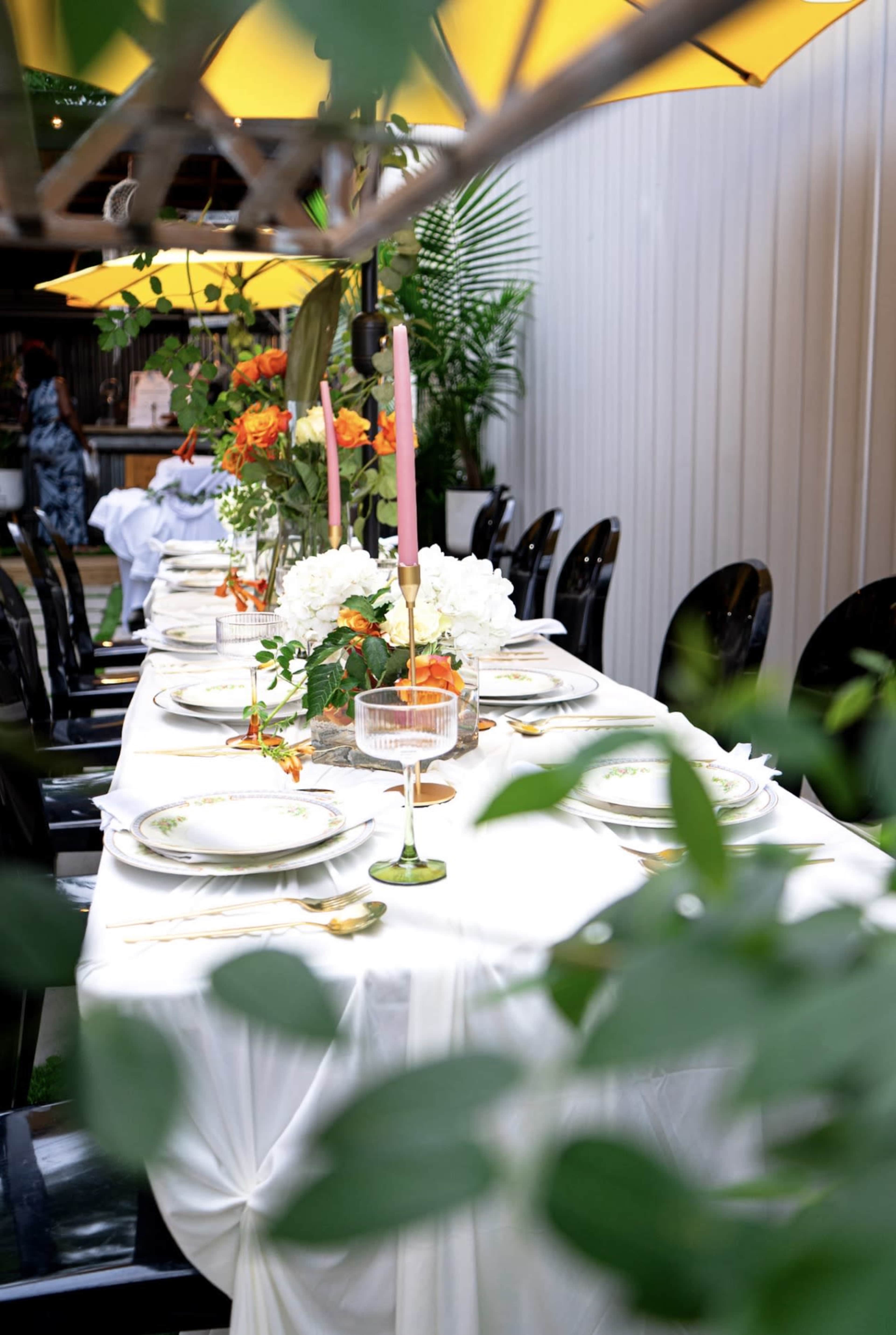 A long, elegantly set dining table with white tablecloth, plates, candle holders, and floral arrangements is placed outdoors under yellow umbrellas.