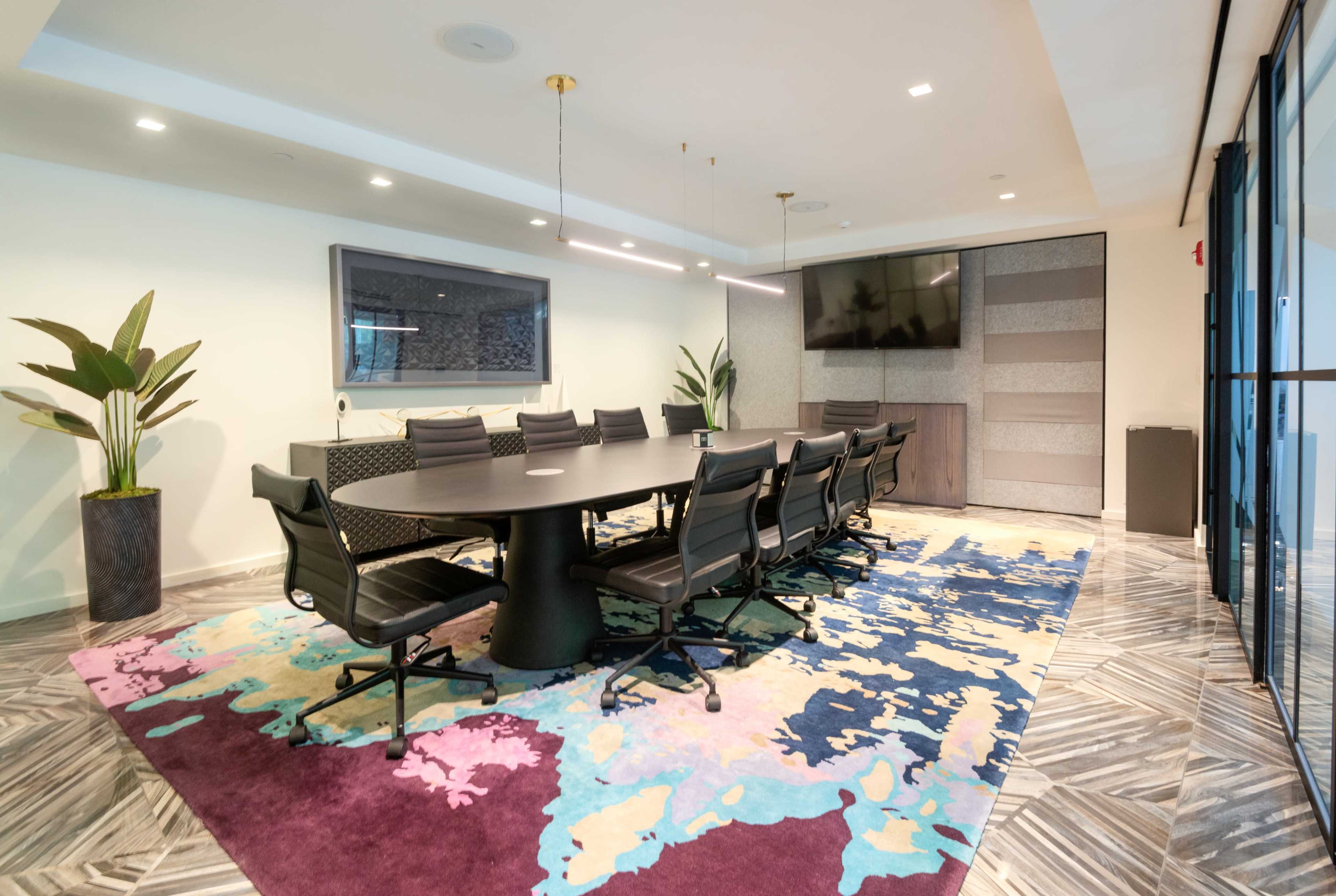 The image shows a modern conference room with a large table surrounded by black chairs, a patterned rug, and a wall-mounted TV.