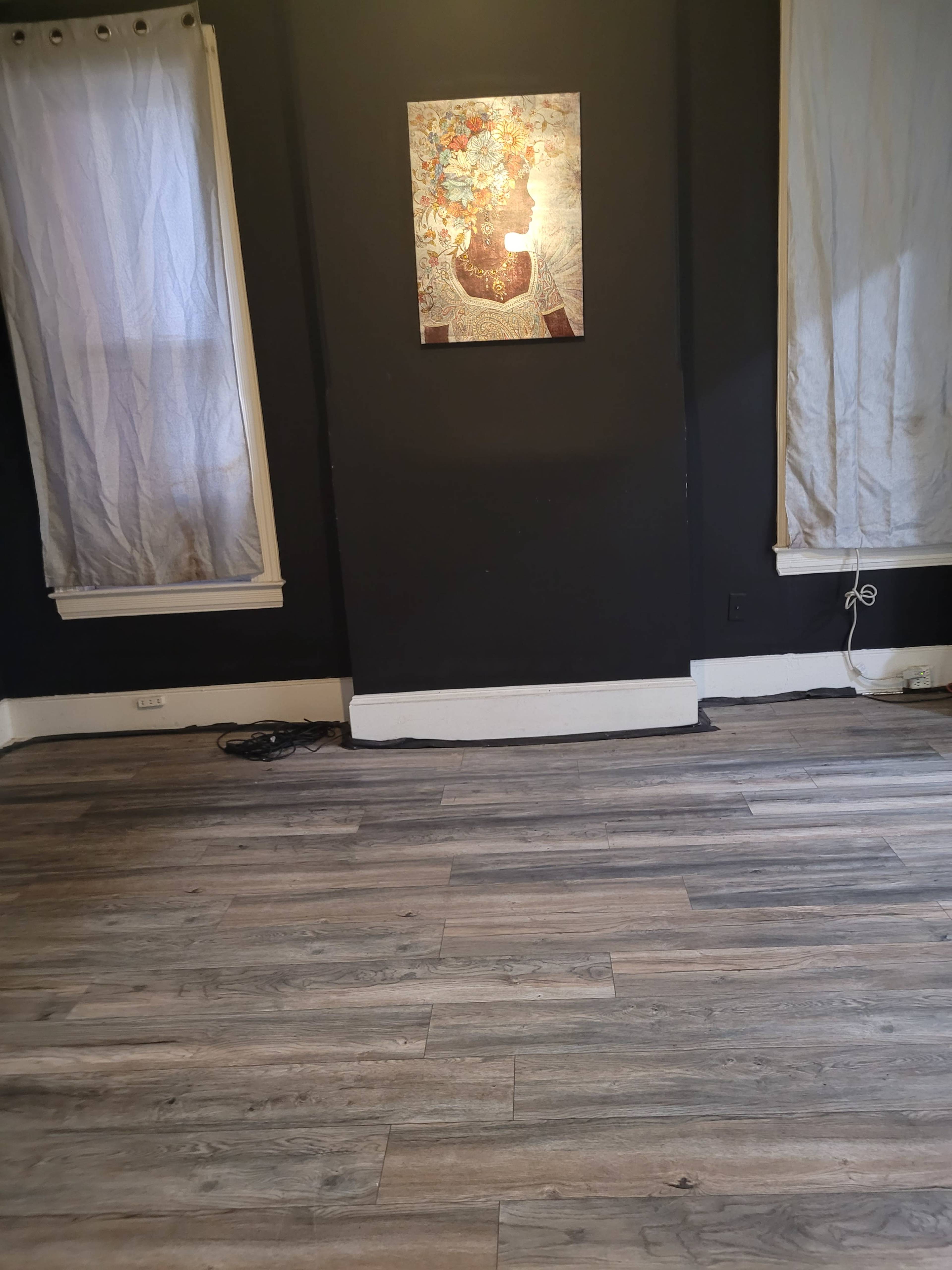The image shows a sparsely furnished room with wooden flooring, two white curtains hanging on the windows, and a framed artwork mounted on a dark wall.