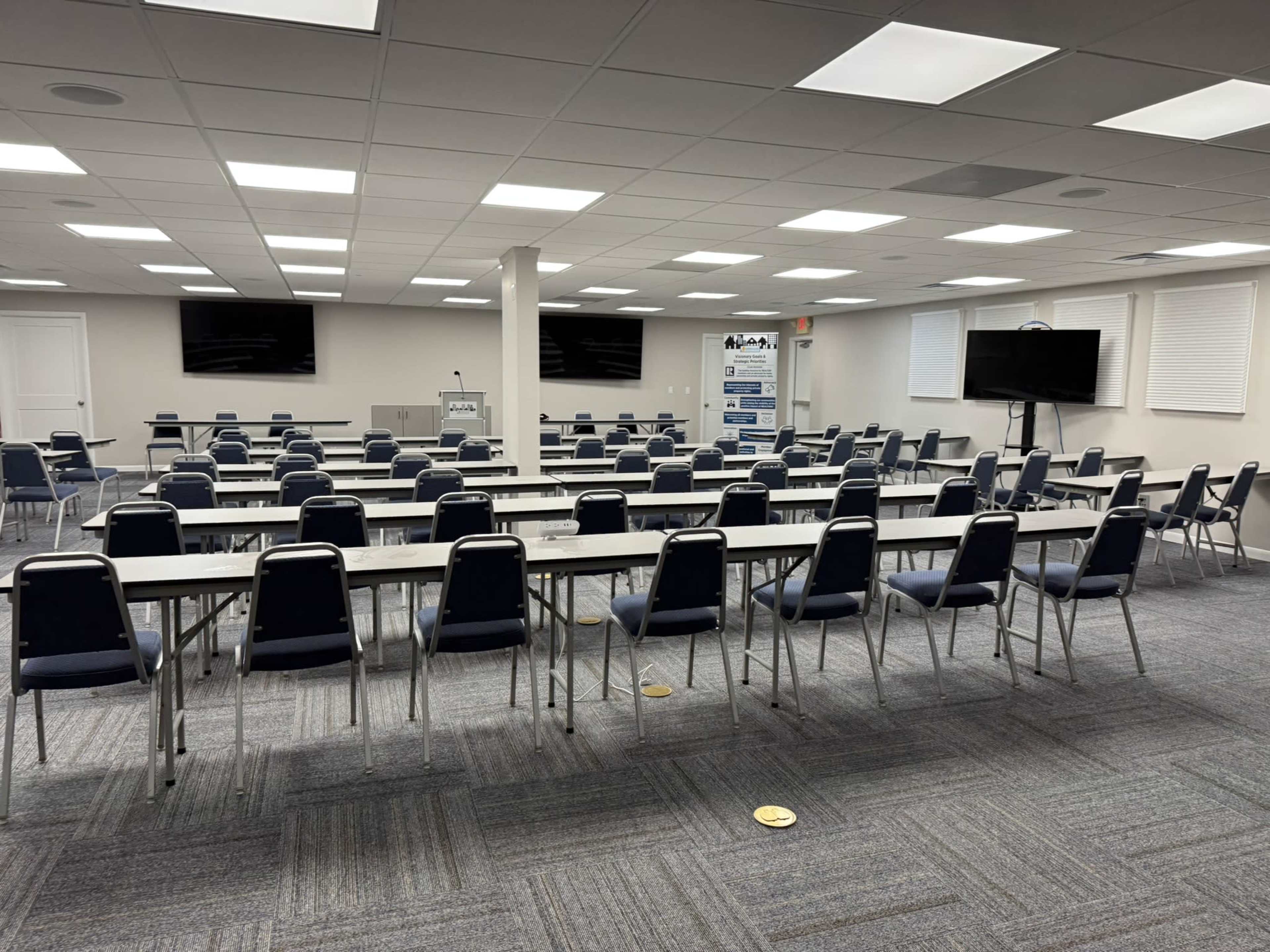 The image shows a conference room set up with multiple rows of tables and chairs, along with two monitors and a display board at the front.