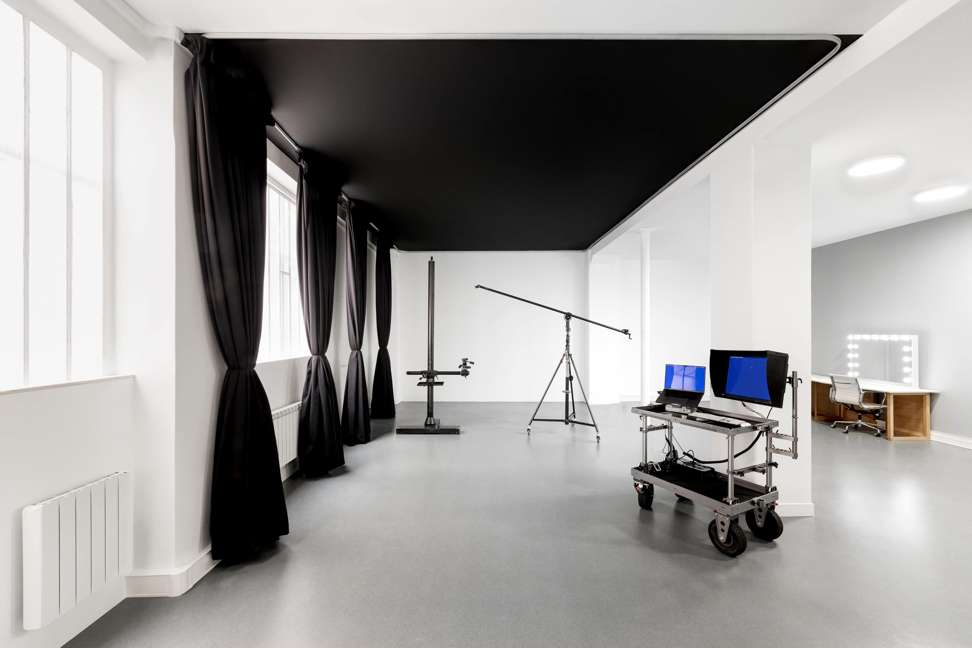 A spacious, minimalistic studio with black curtains, a lighting stand, a computer workstation on wheels, and a vanity area with illuminated mirrors.