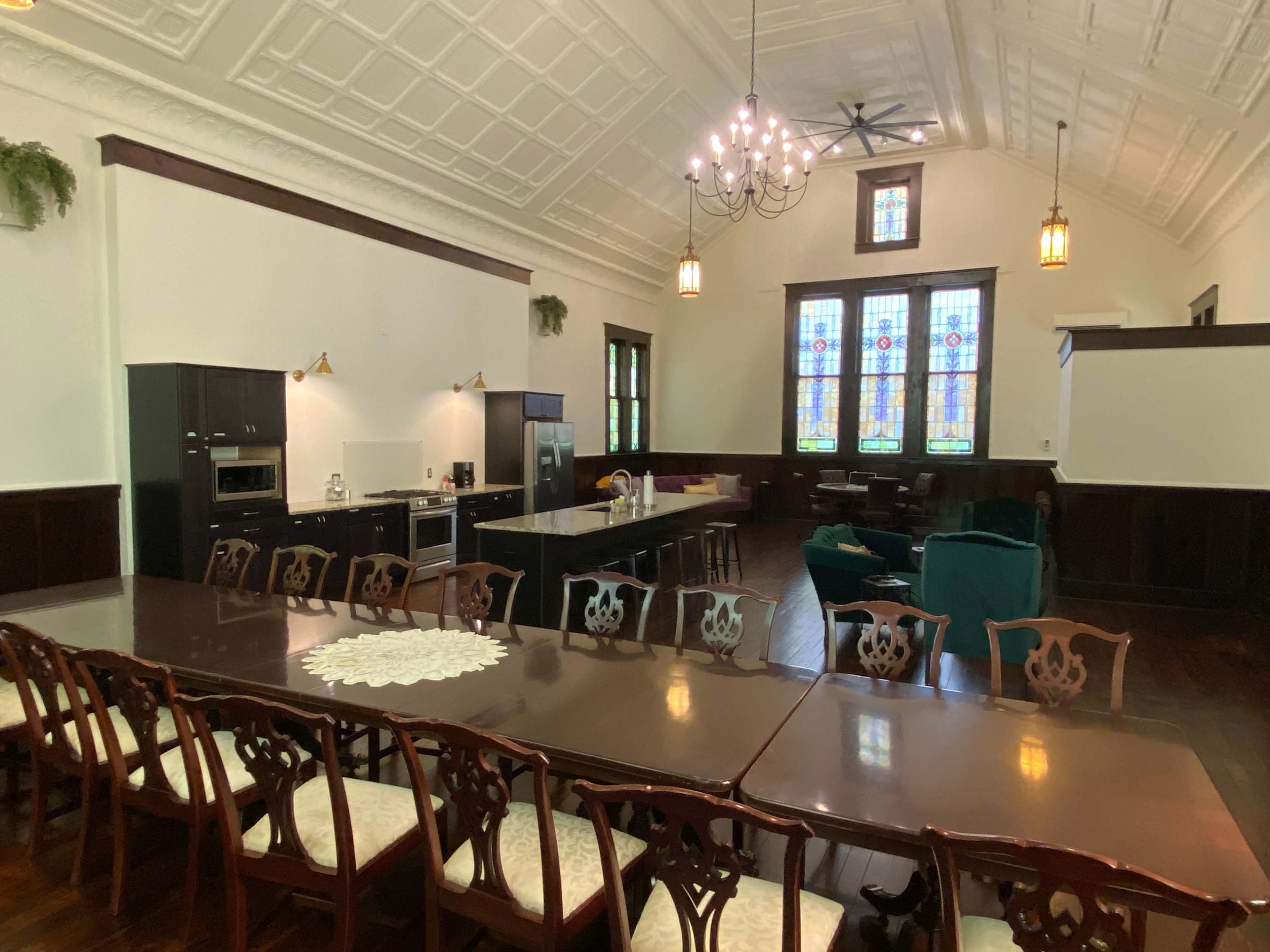 The image shows a spacious room with a long dining table, a modern kitchen area, and large stained glass windows.