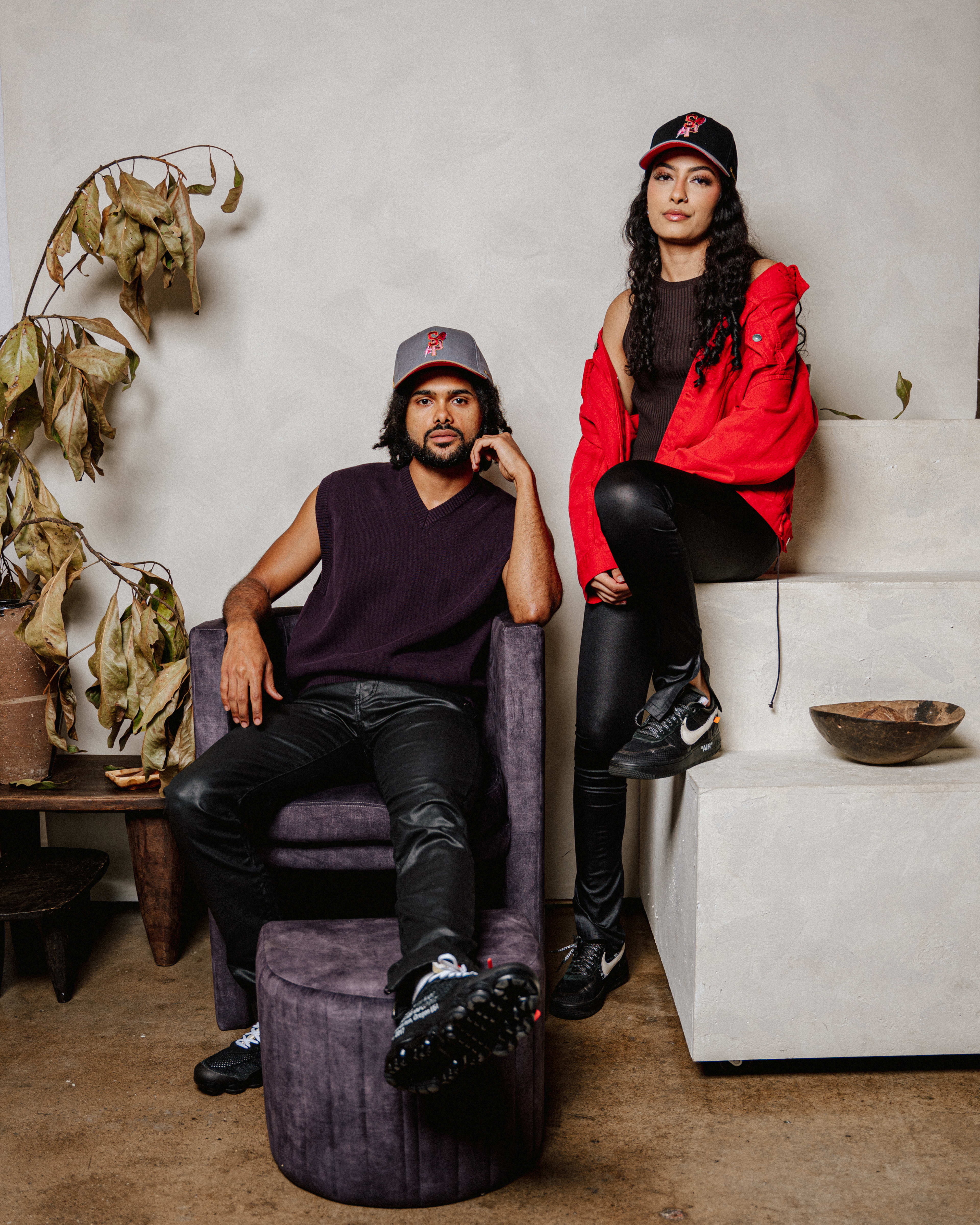 A rustic photoshoot with a man and a woman posing in baseball hats and black leather pants.
