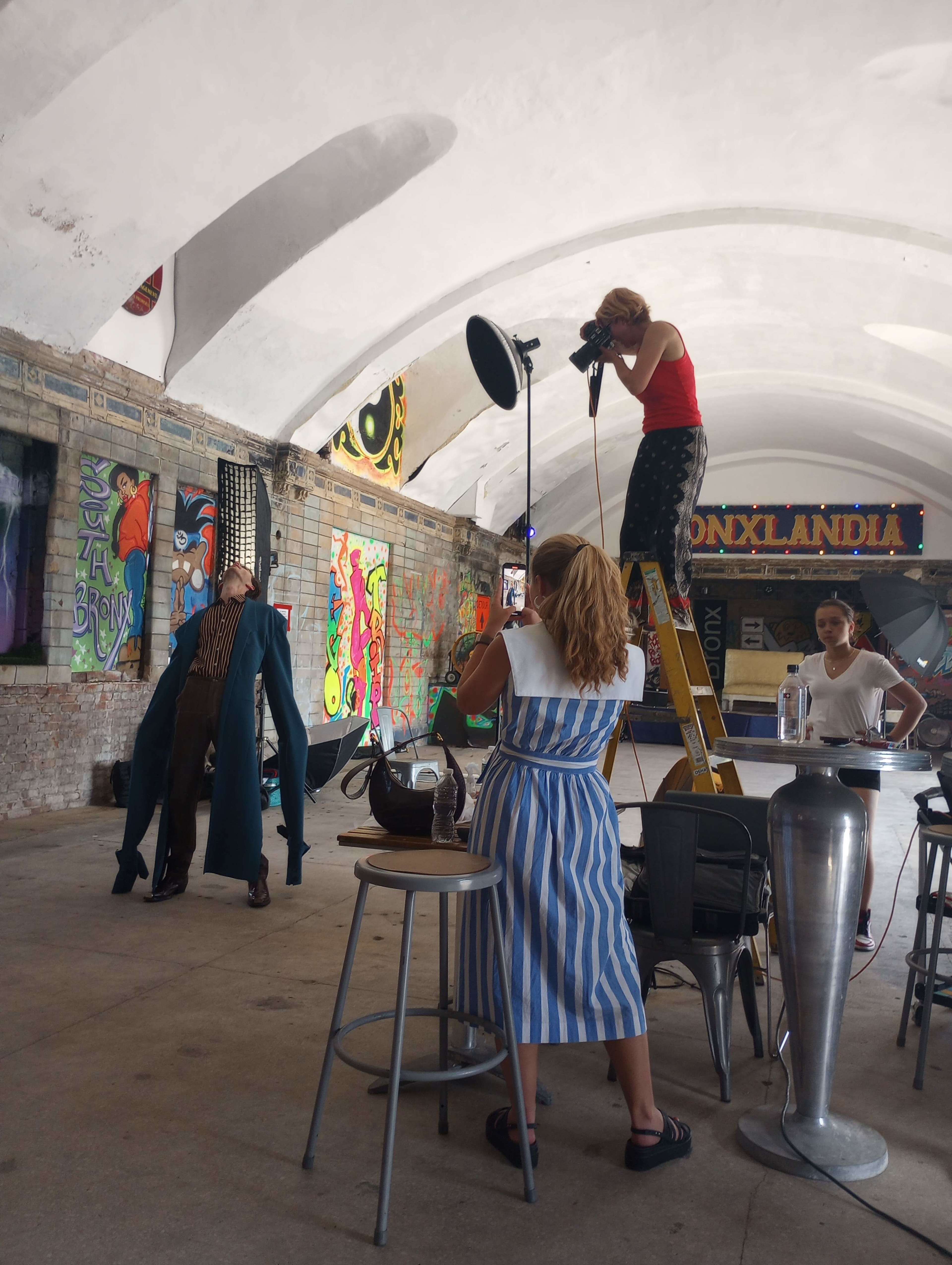 A fashion photoshoot is taking place indoors, featuring a model in a striking outfit and a photographer on a ladder adjusting lights.