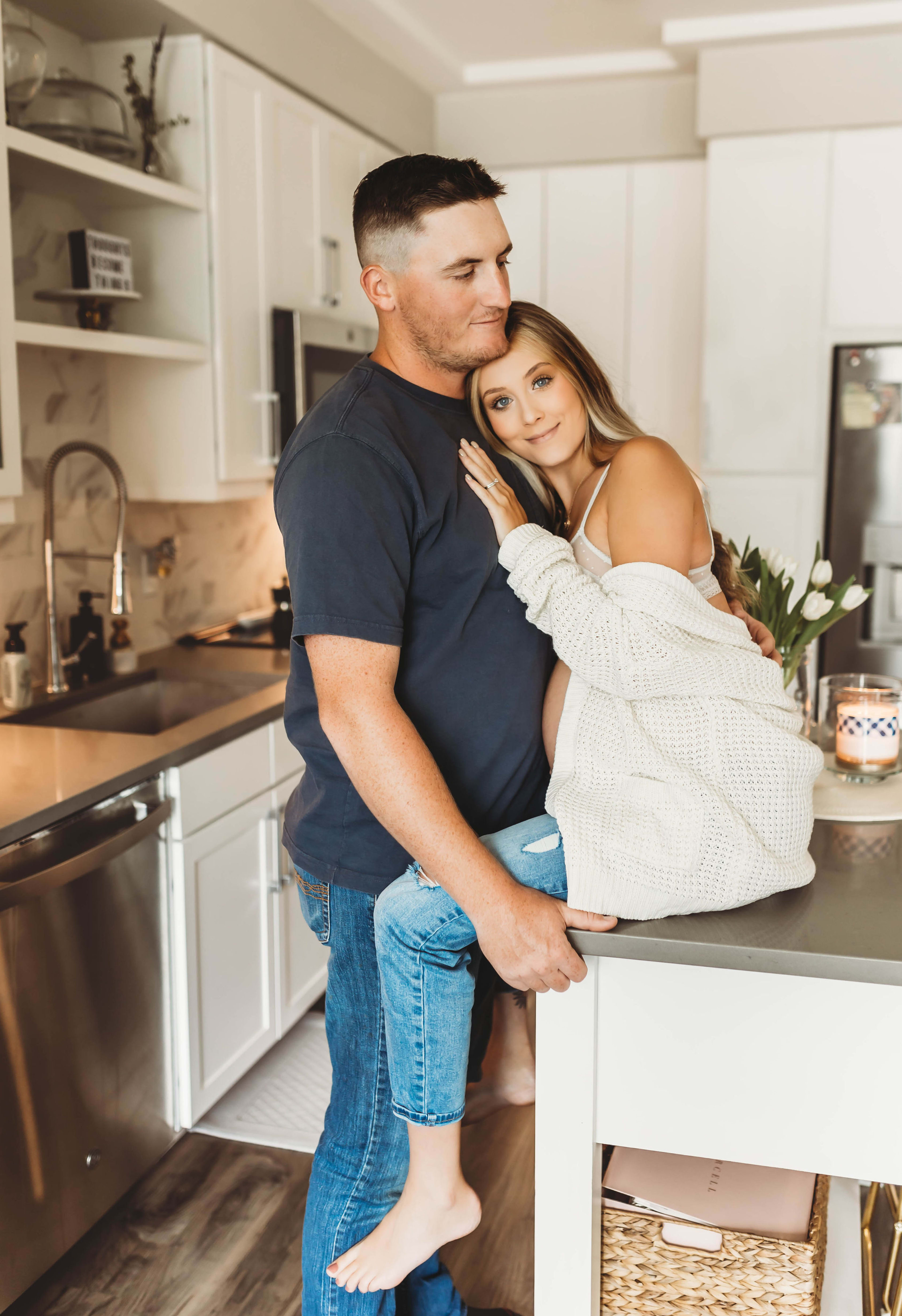 Una mujer embarazada y su marido abrazados en su cocina blanca durante una sesión de fotos de maternidad.