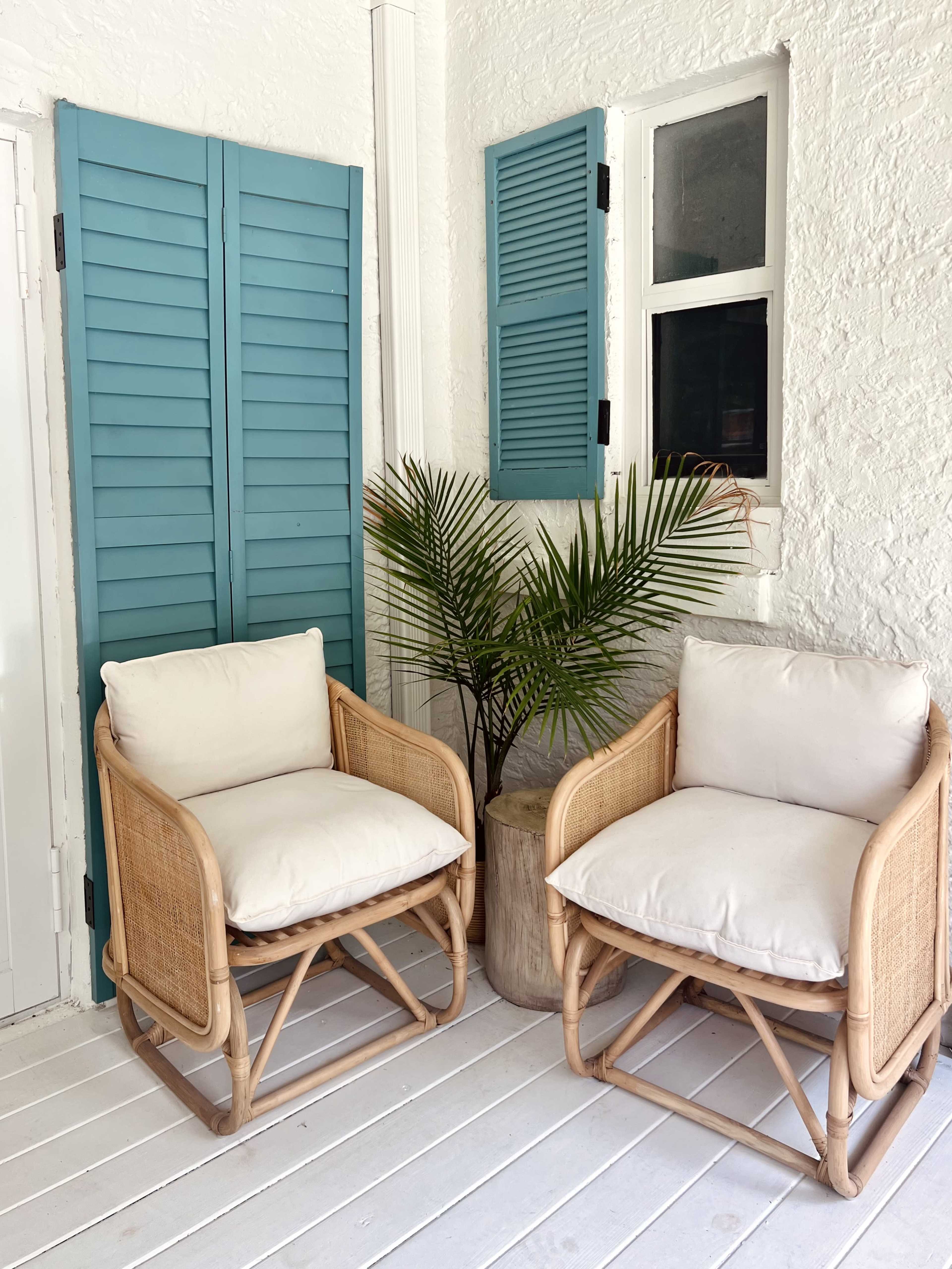 Two rattan chairs with white cushions, positioned next to a potted plant and blue-shuttered windows in a white-walled interior space.