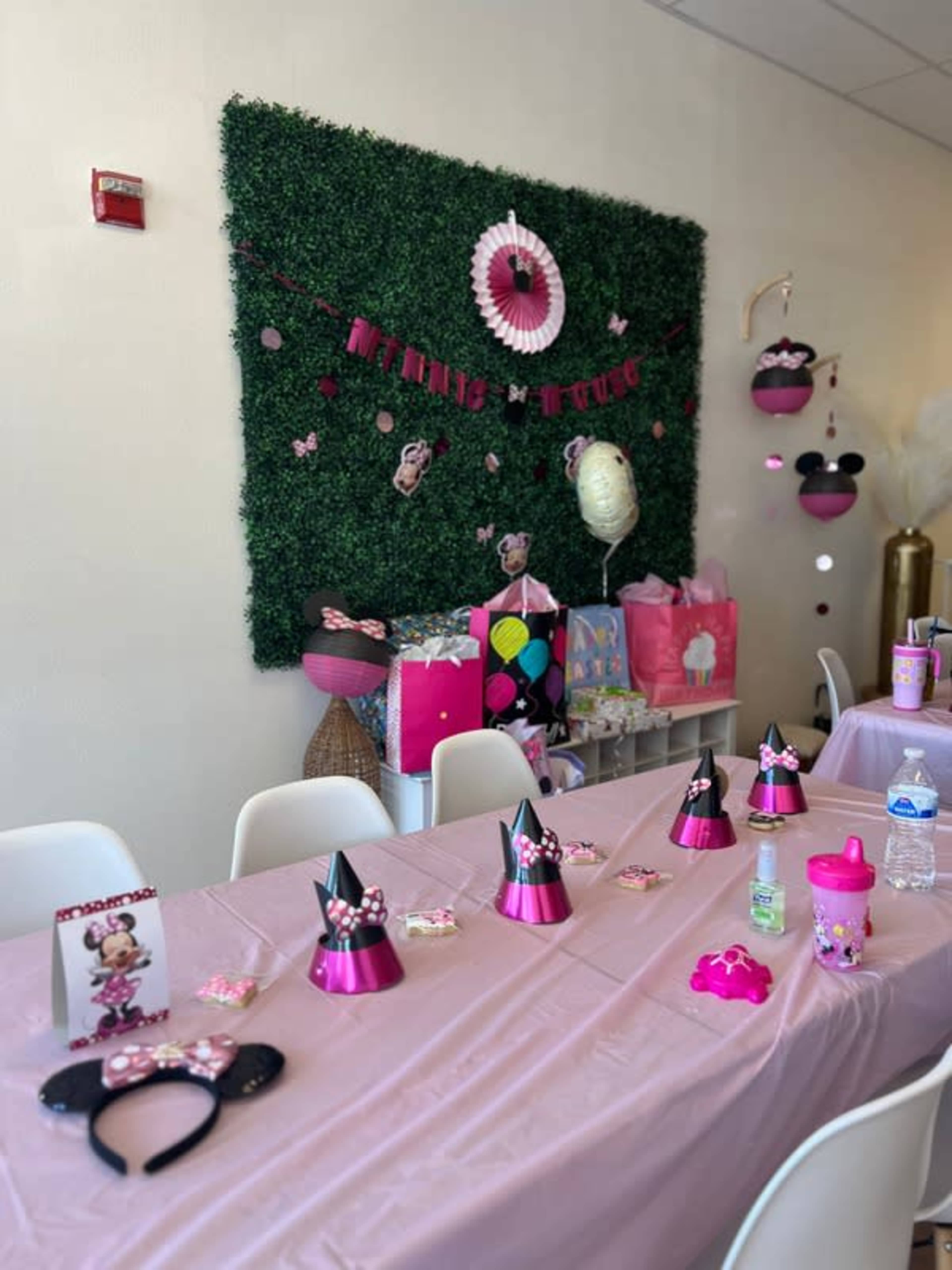 A pink-themed birthday party setup features a table adorned with party hats and decorations, alongside a wall decorated with greenery and balloons.
