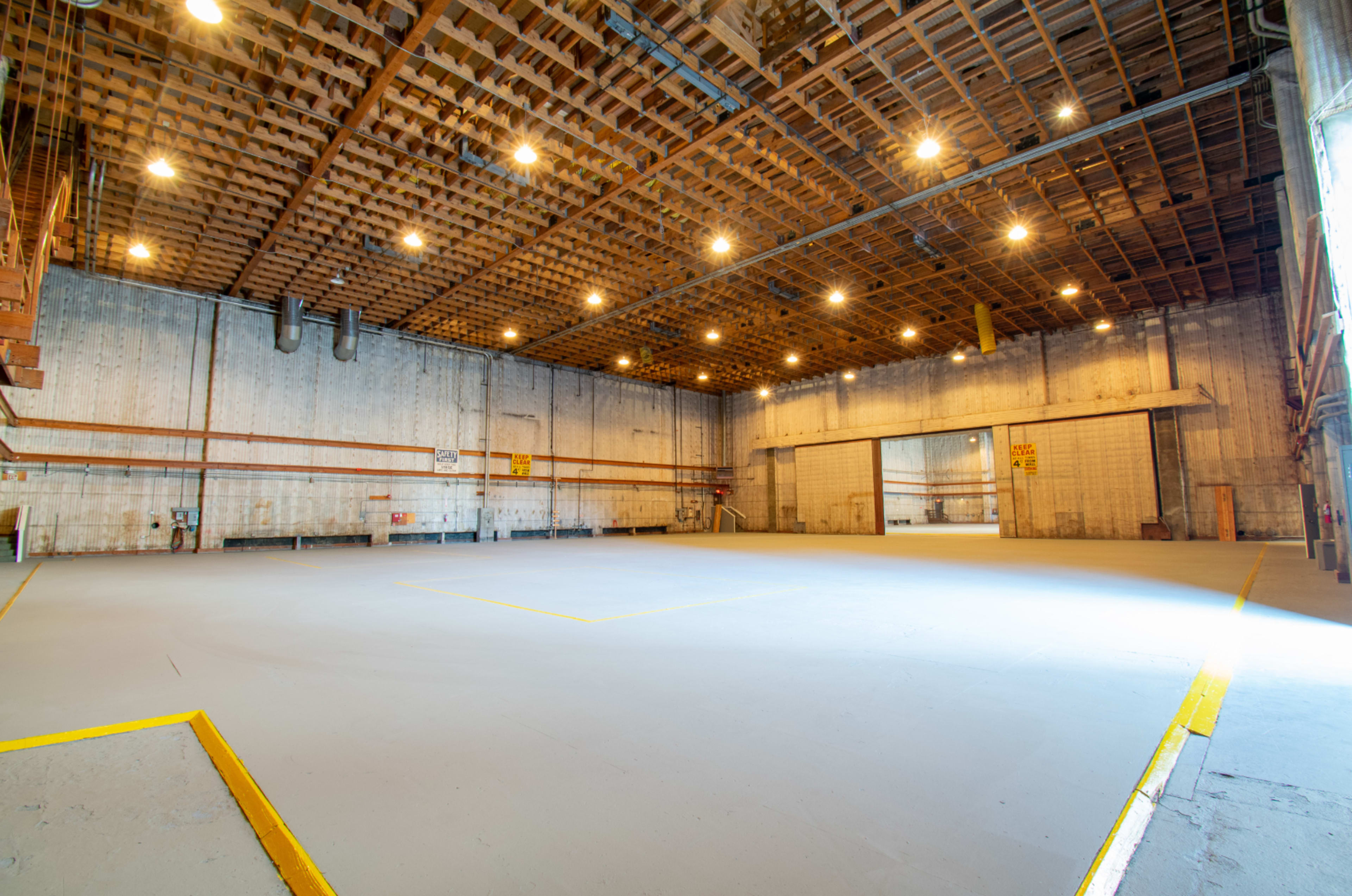 The image shows an empty warehouse interior with high ceilings, overhead lighting, and large open spaces.