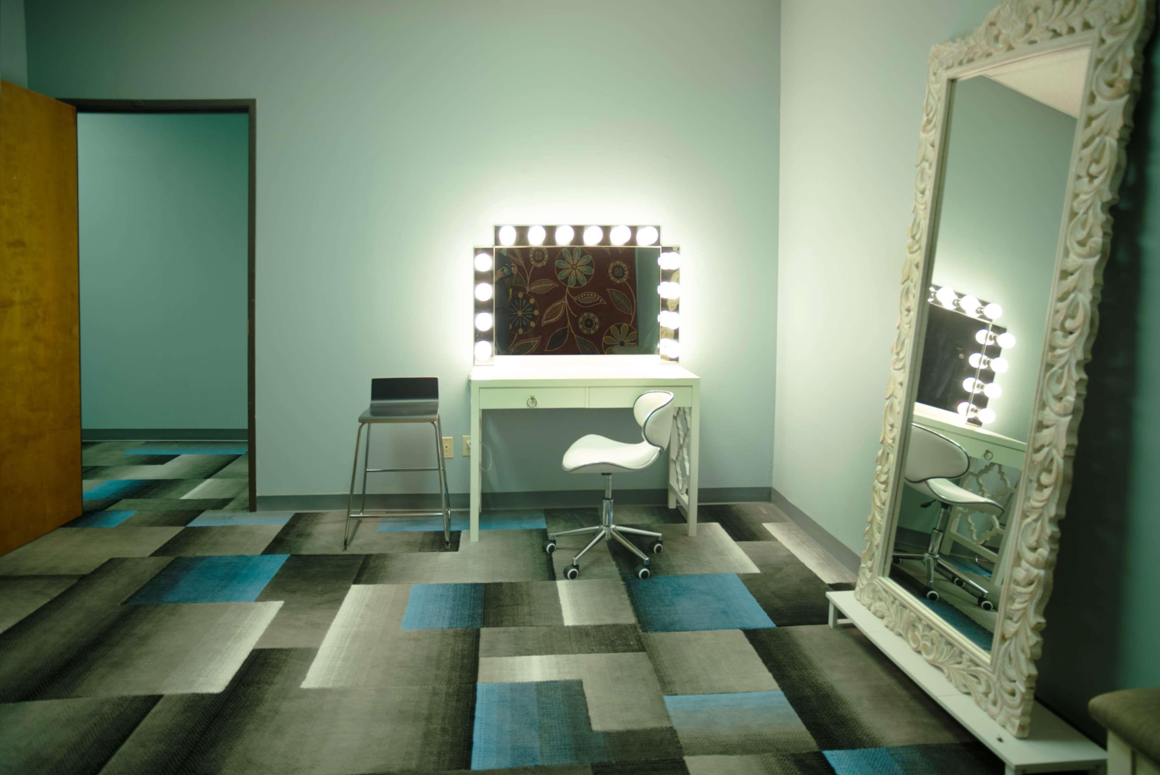 The image shows a brightly lit vanity area with a mirror, a chair, and a large decorative mirror against a wall in a room with blue-green walls and a patterned carpet.