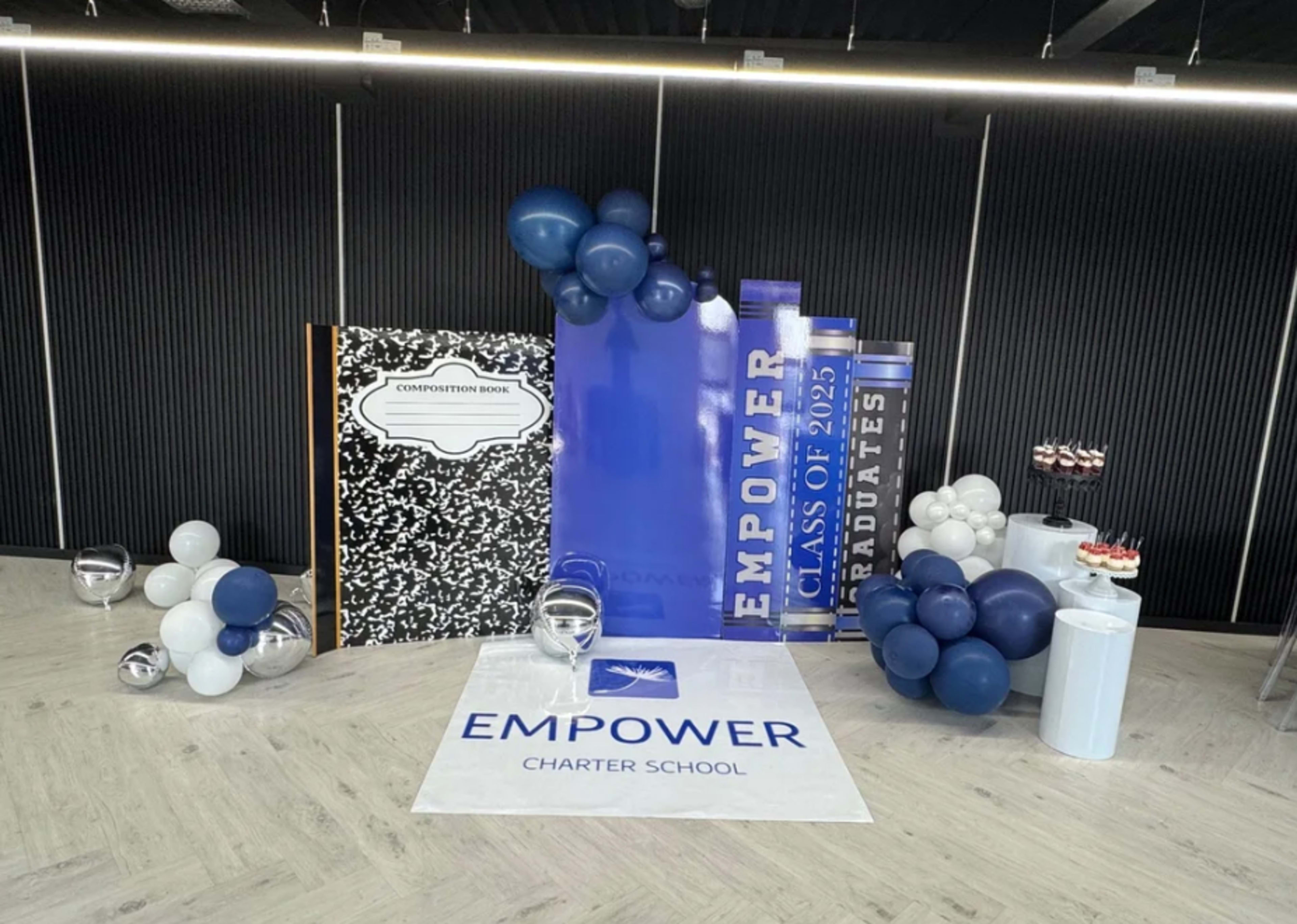The image shows a decorated area for a graduation event at Empower Charter School, featuring a large faux composition book, blue balloons, a sign for the class of 2025, and a display of cupcakes.