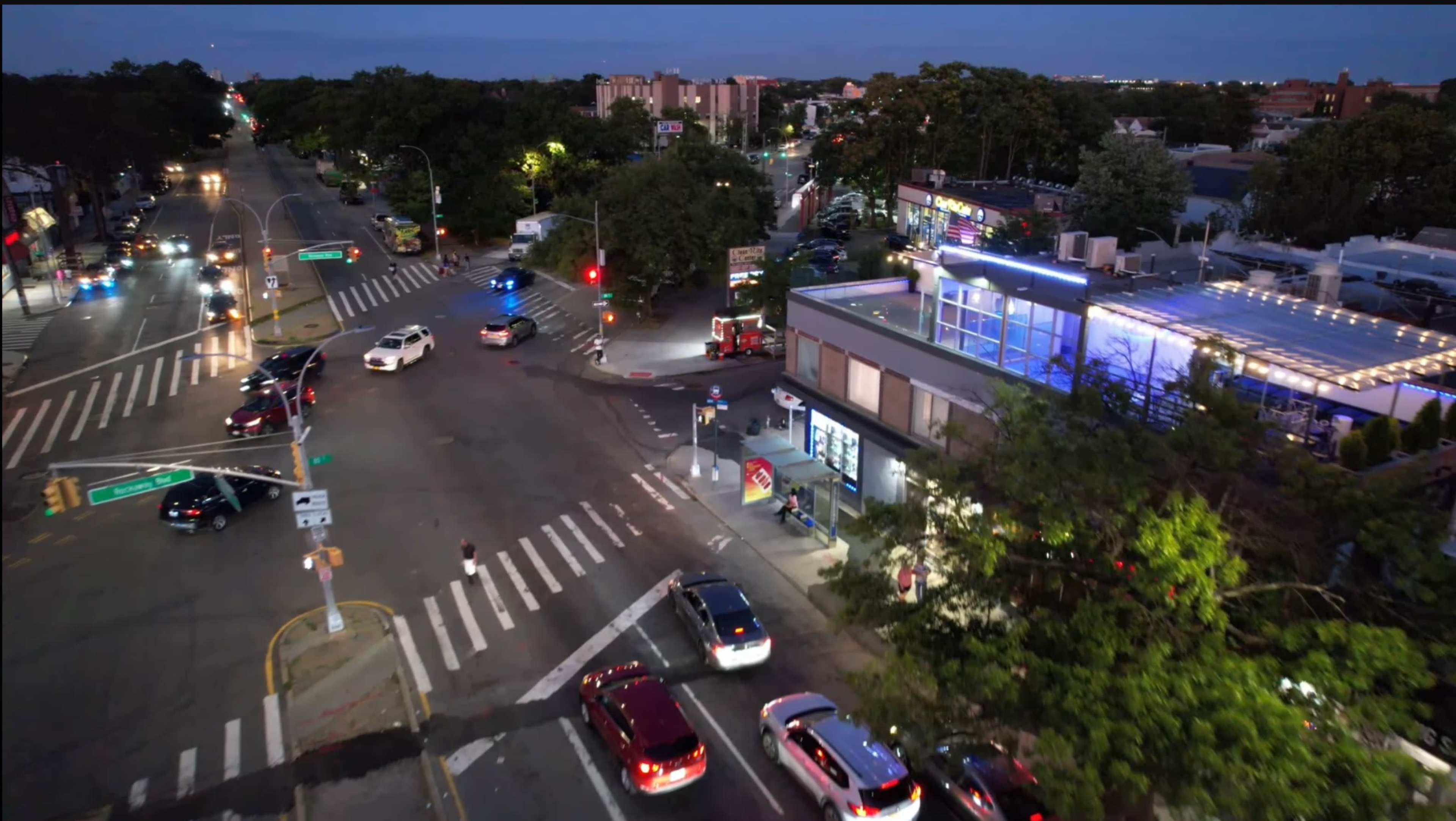 The scene shows a busy urban intersection at dusk, with cars passing through traffic lights and a nearby building illuminated by blue lights.
