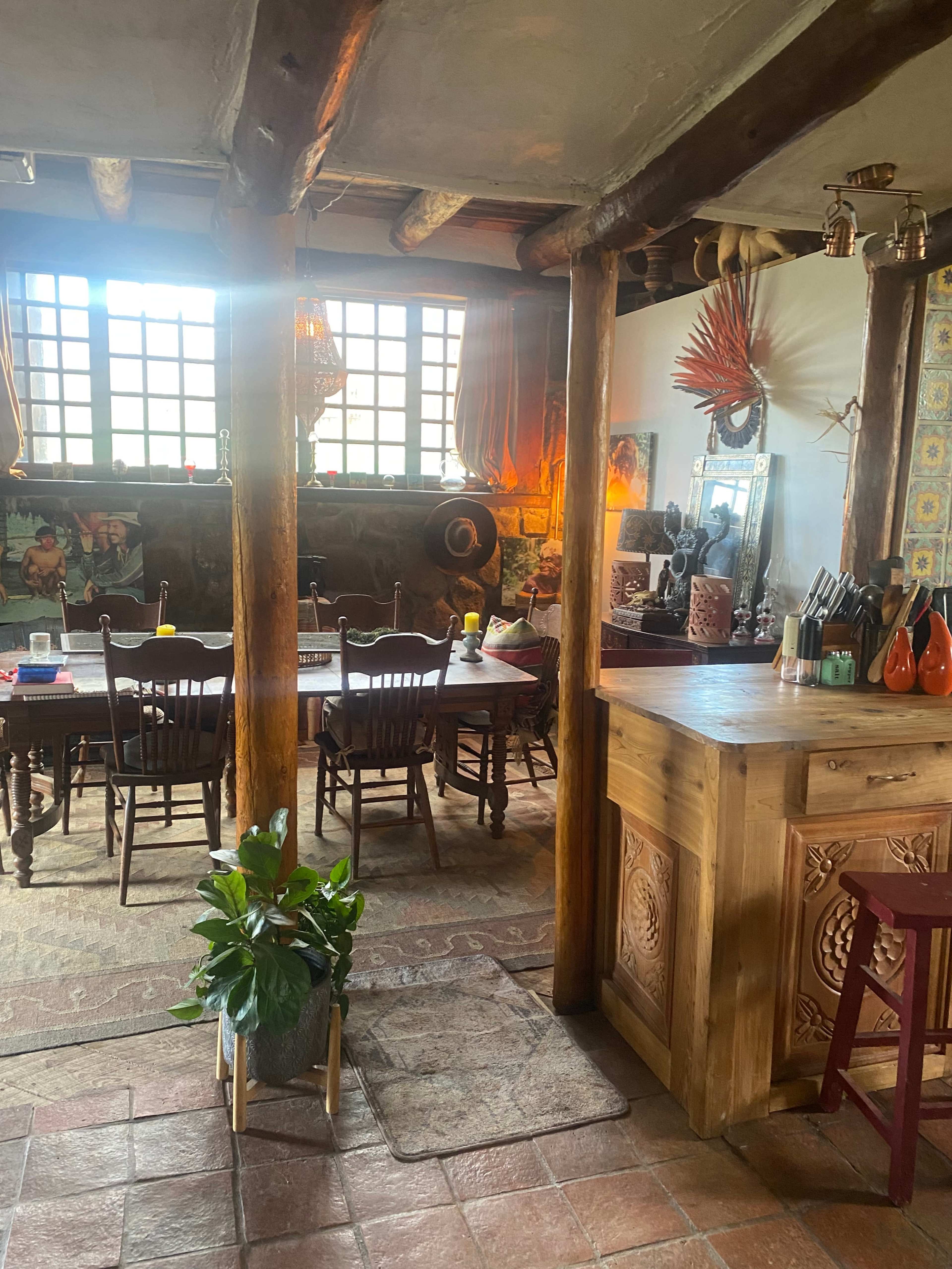 The image shows a rustic dining area with wooden beams, a long table set for several guests, and a view of a kitchen counter with various utensils and a potted plant.