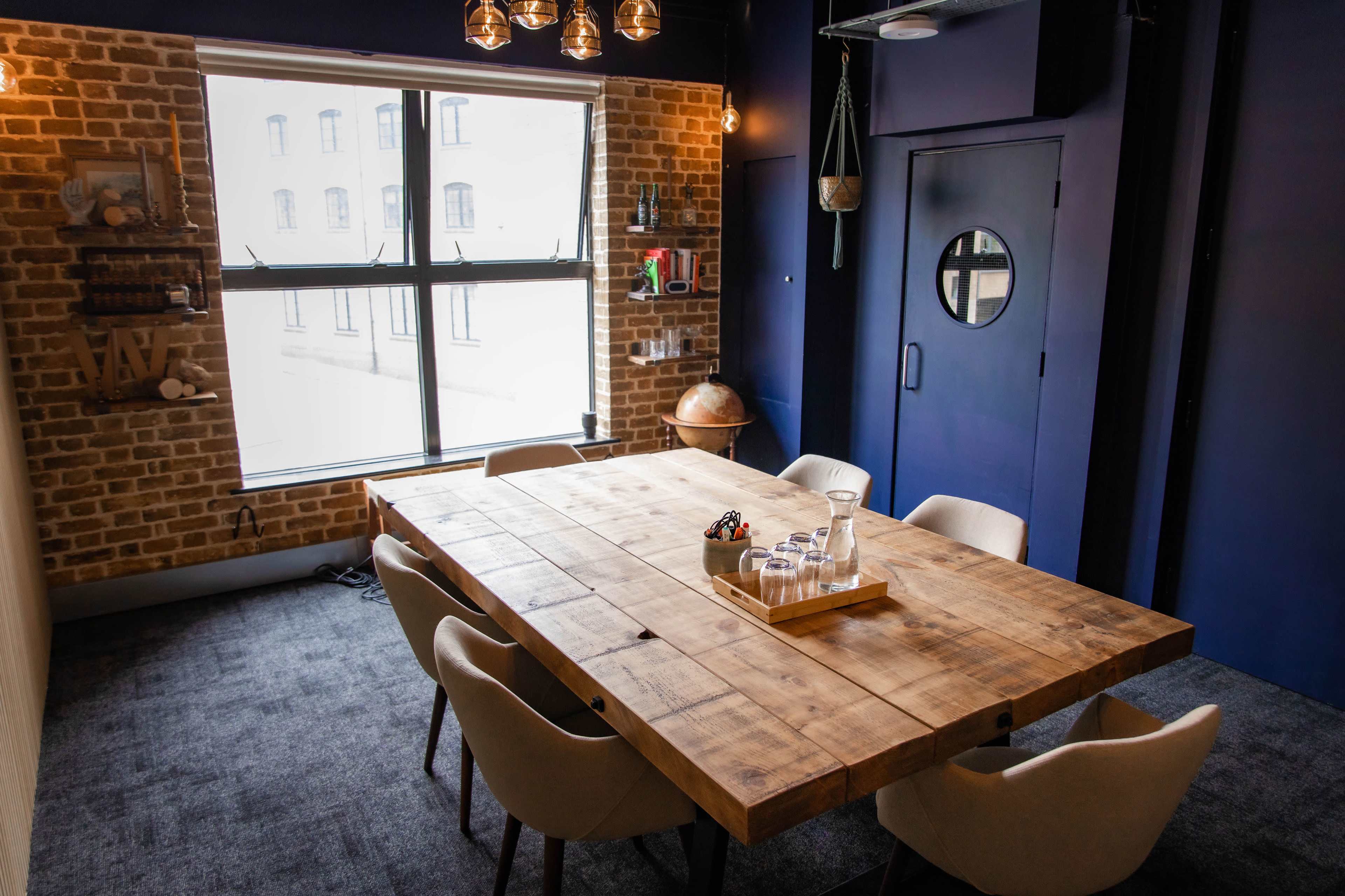 A meeting room with a large wooden table surrounded by beige chairs, a window with multiple panes, and exposed brick walls.