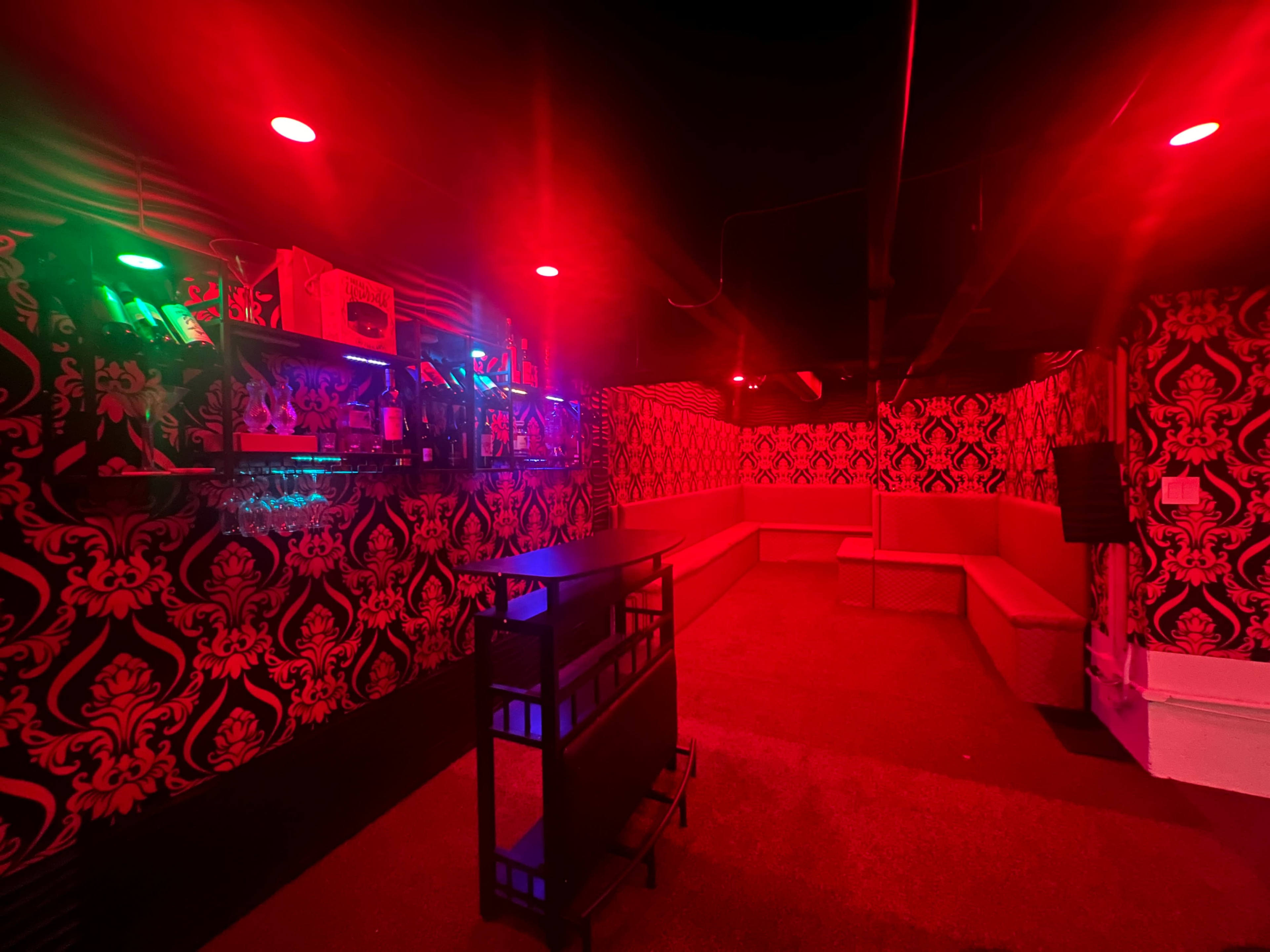The image shows a dimly lit room with red and green lighting, featuring patterned wallpaper and a bar area against one wall.