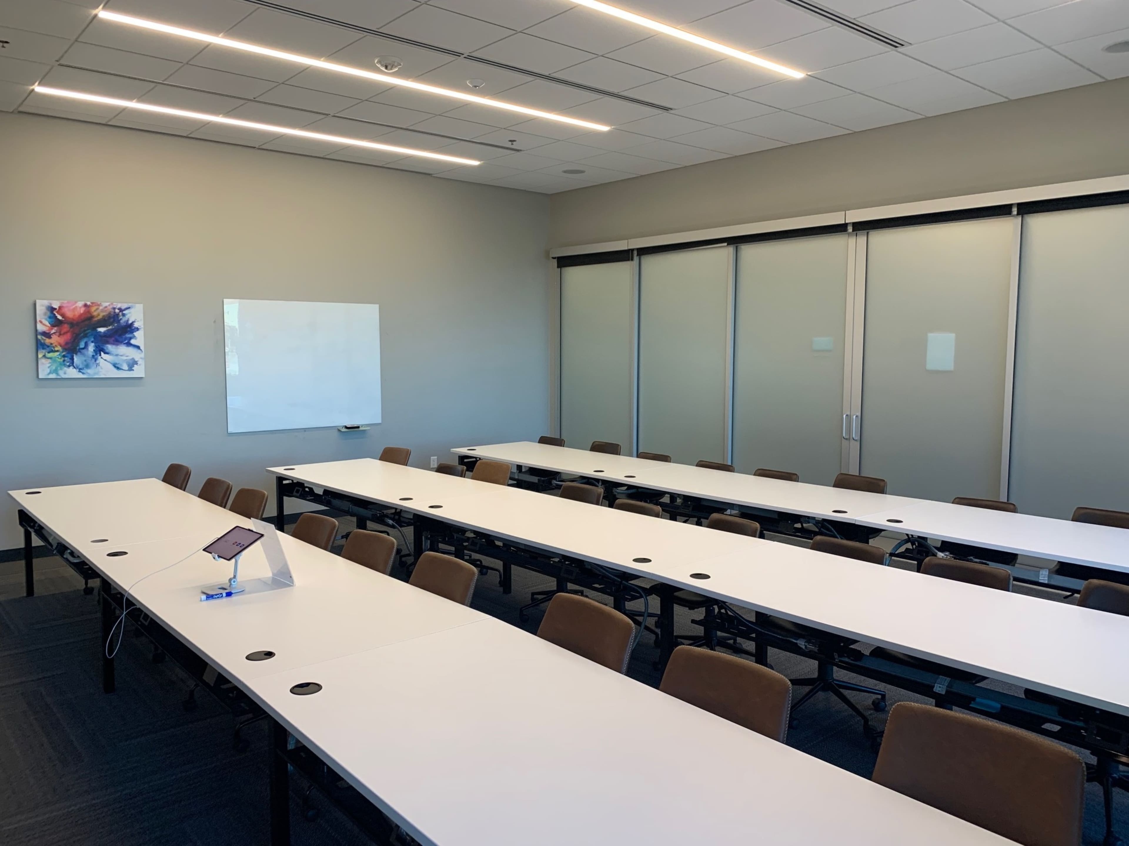 A spacious meeting room is set up with multiple rows of white tables and brown chairs, featuring a whiteboard and a colorful artwork on the wall.