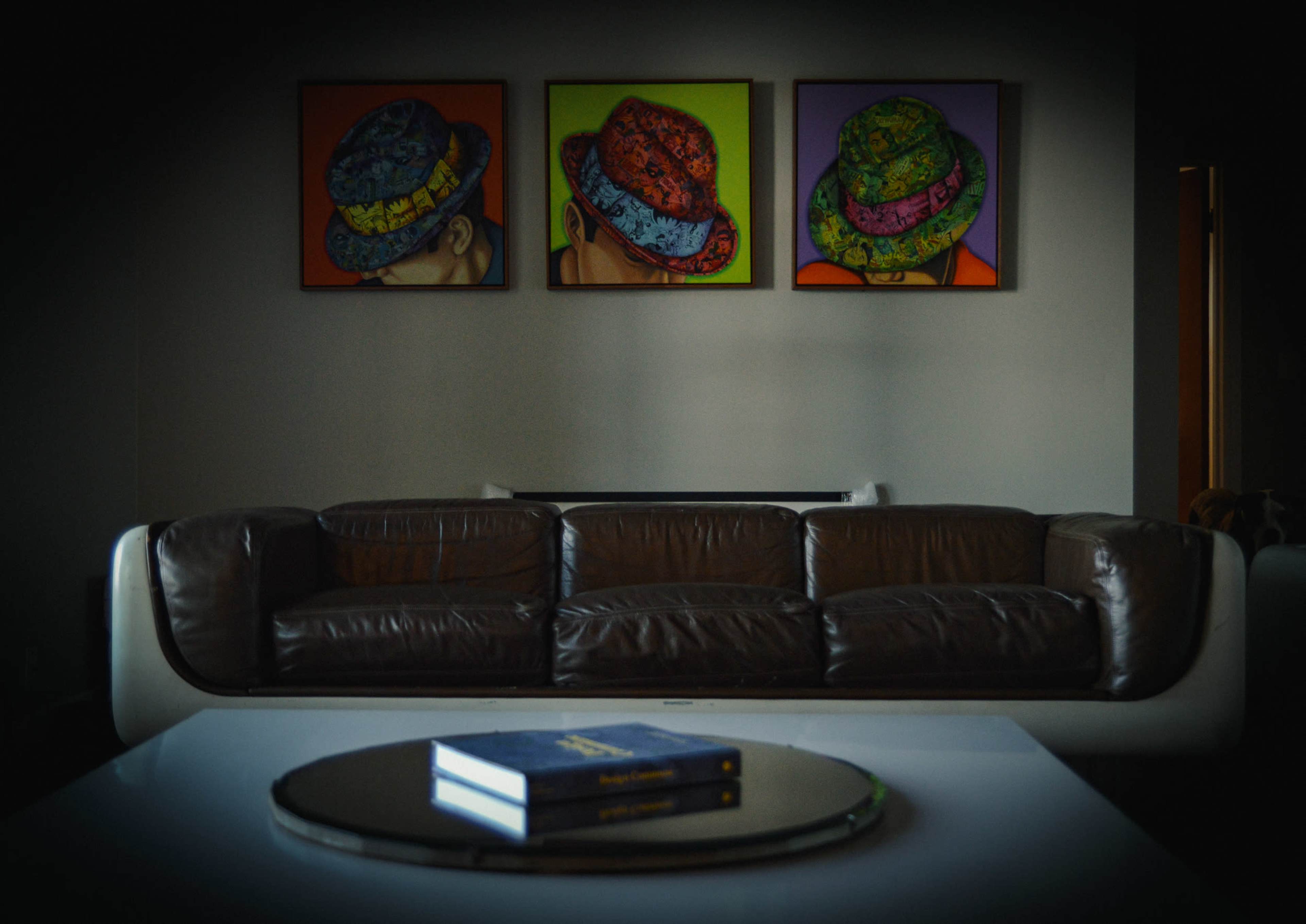 A brown leather sofa faces a coffee table with two books, while colorful hat-themed artworks hang on the wall behind it.