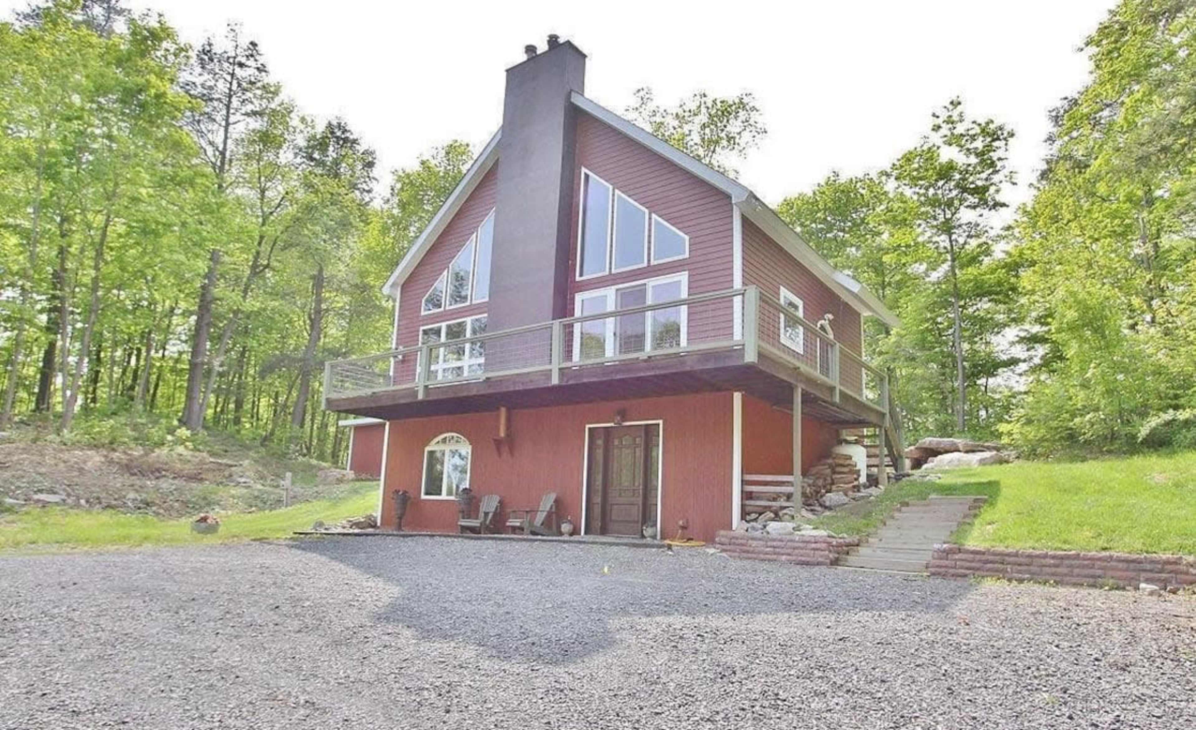 A two-story red house with large windows and a balcony is situated on a gravel driveway surrounded by trees.