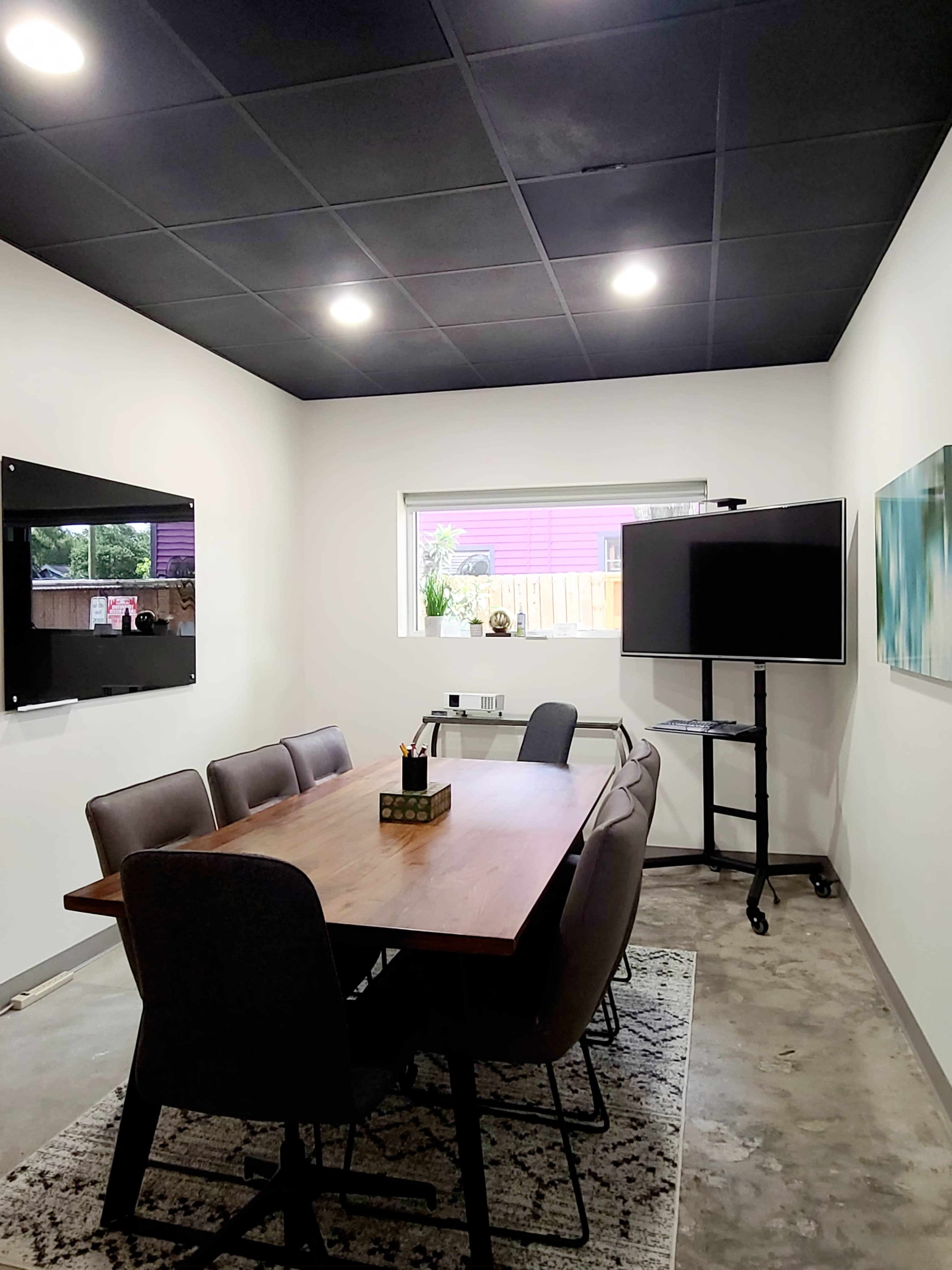 A modern conference room features a large wooden table surrounded by eight chairs, with two wall-mounted screens and a mobile display in the corner.