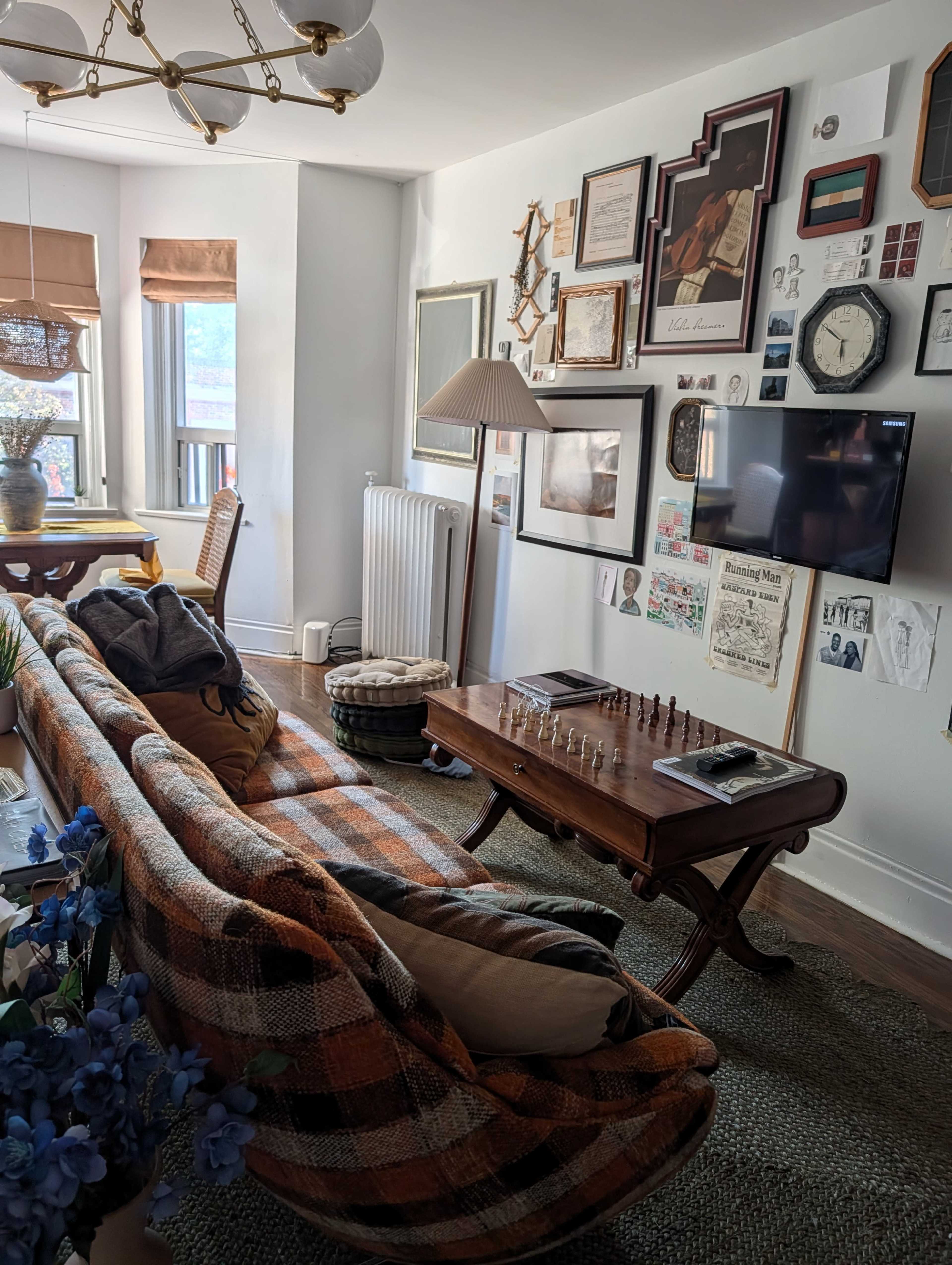 A plaid sofa faces a coffee table with a chess set, surrounded by an assortment of framed pictures and decor on the walls of a light-filled living room.