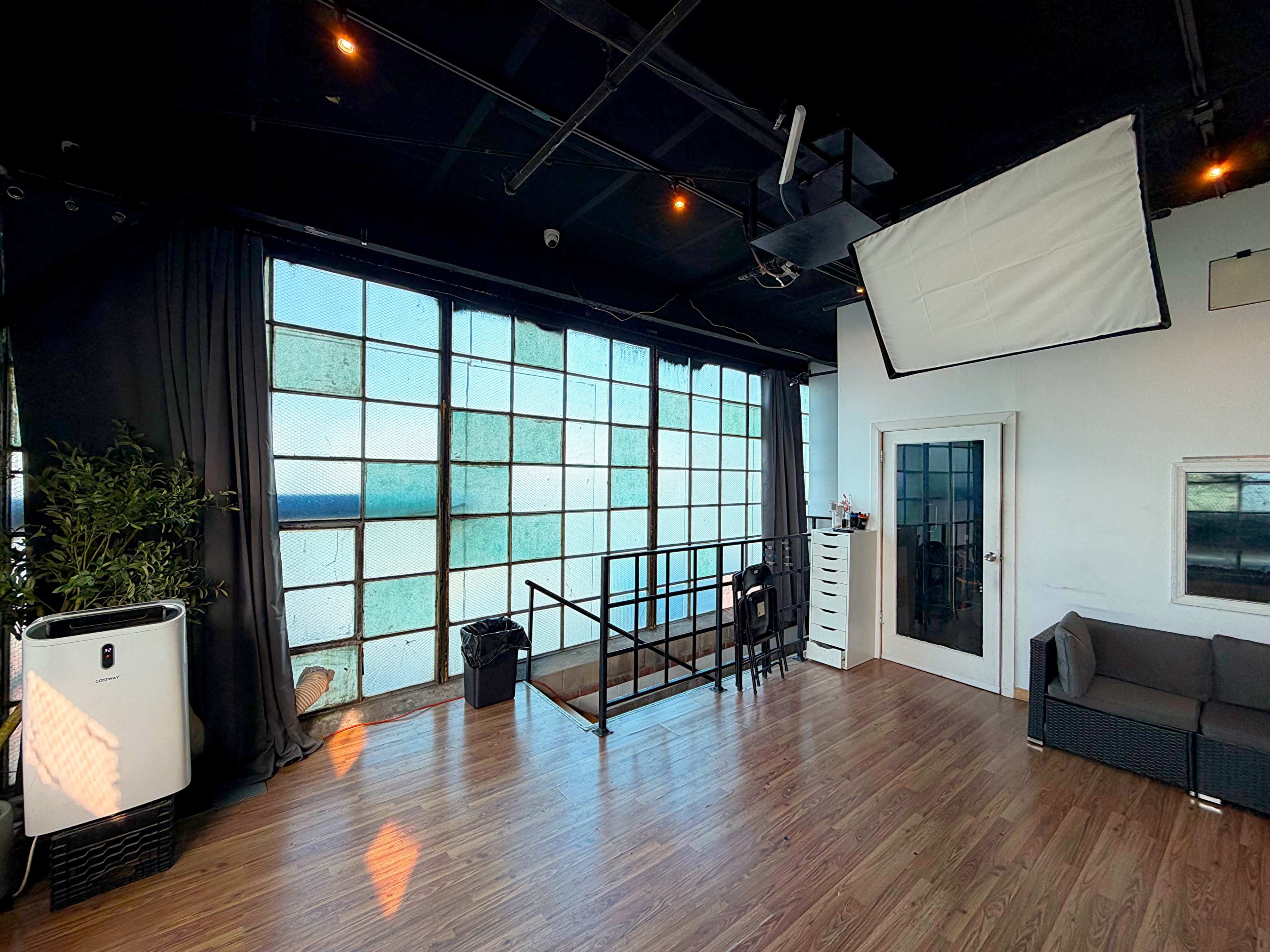The image shows a spacious studio with large glass windows, wooden flooring, and a staircase leading to a lower area, accompanied by minimalist furniture and a plant.
