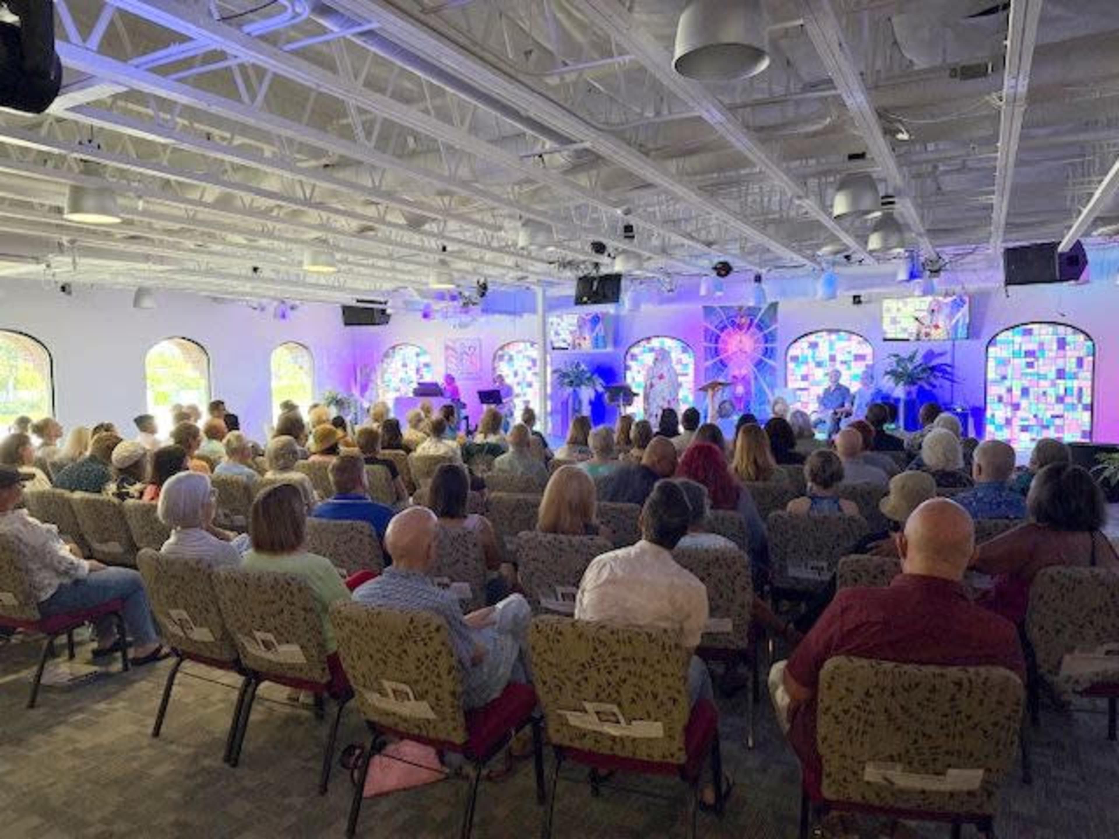 A large audience is seated in a well-lit room with colorful stained glass windows, facing a stage with screens displaying images.